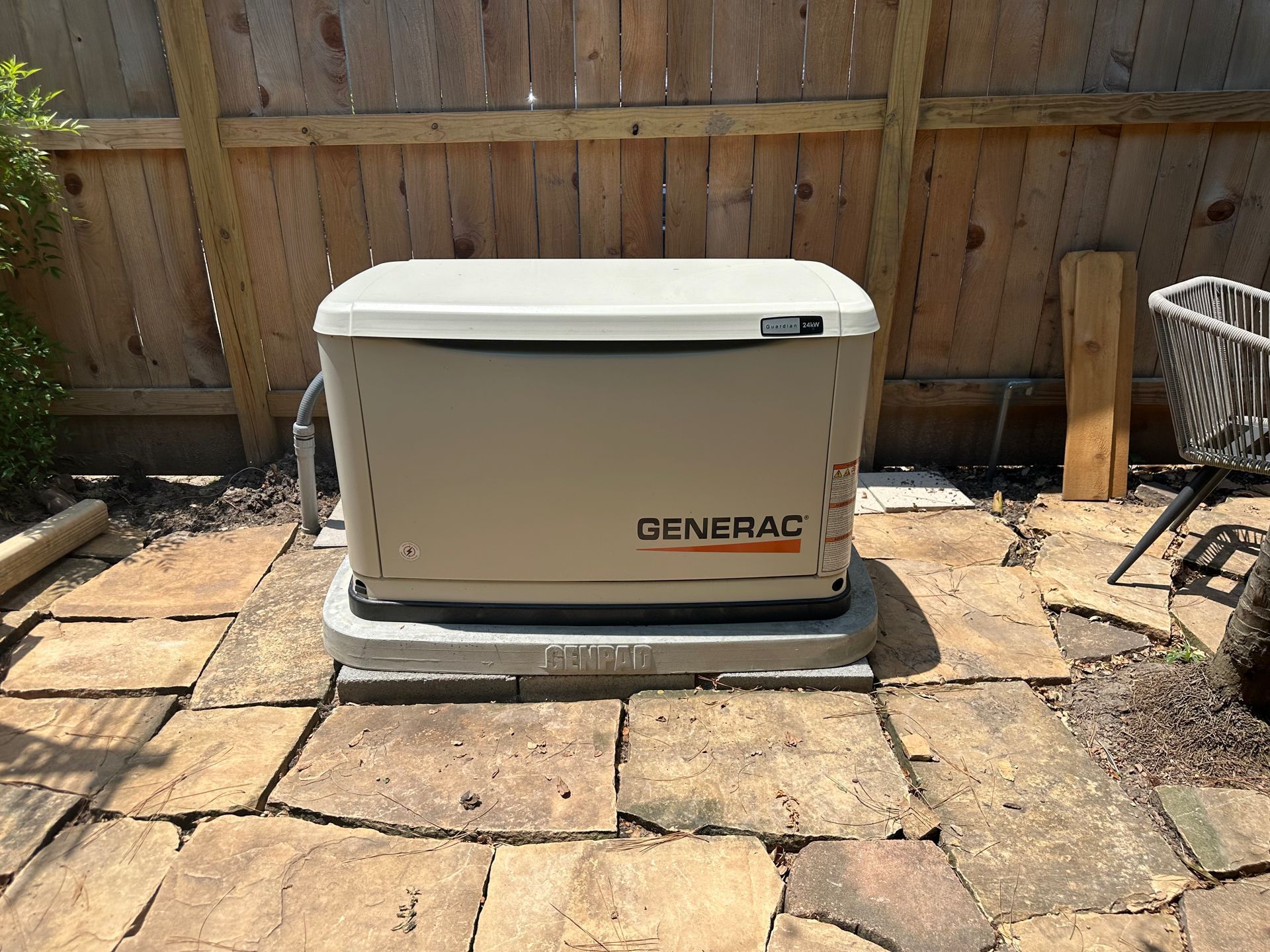 A Generac home generator on a concrete base, in a backyard setting, near a wooden fence.