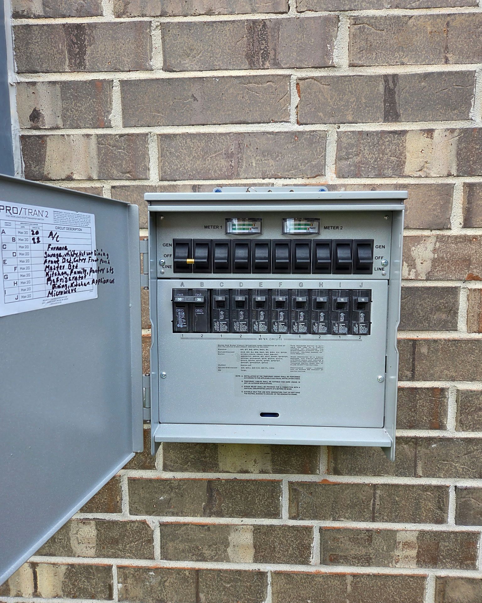 Outdoor electrical panel with open gray door, mounted on a brick wall. Circuit breakers are visible.