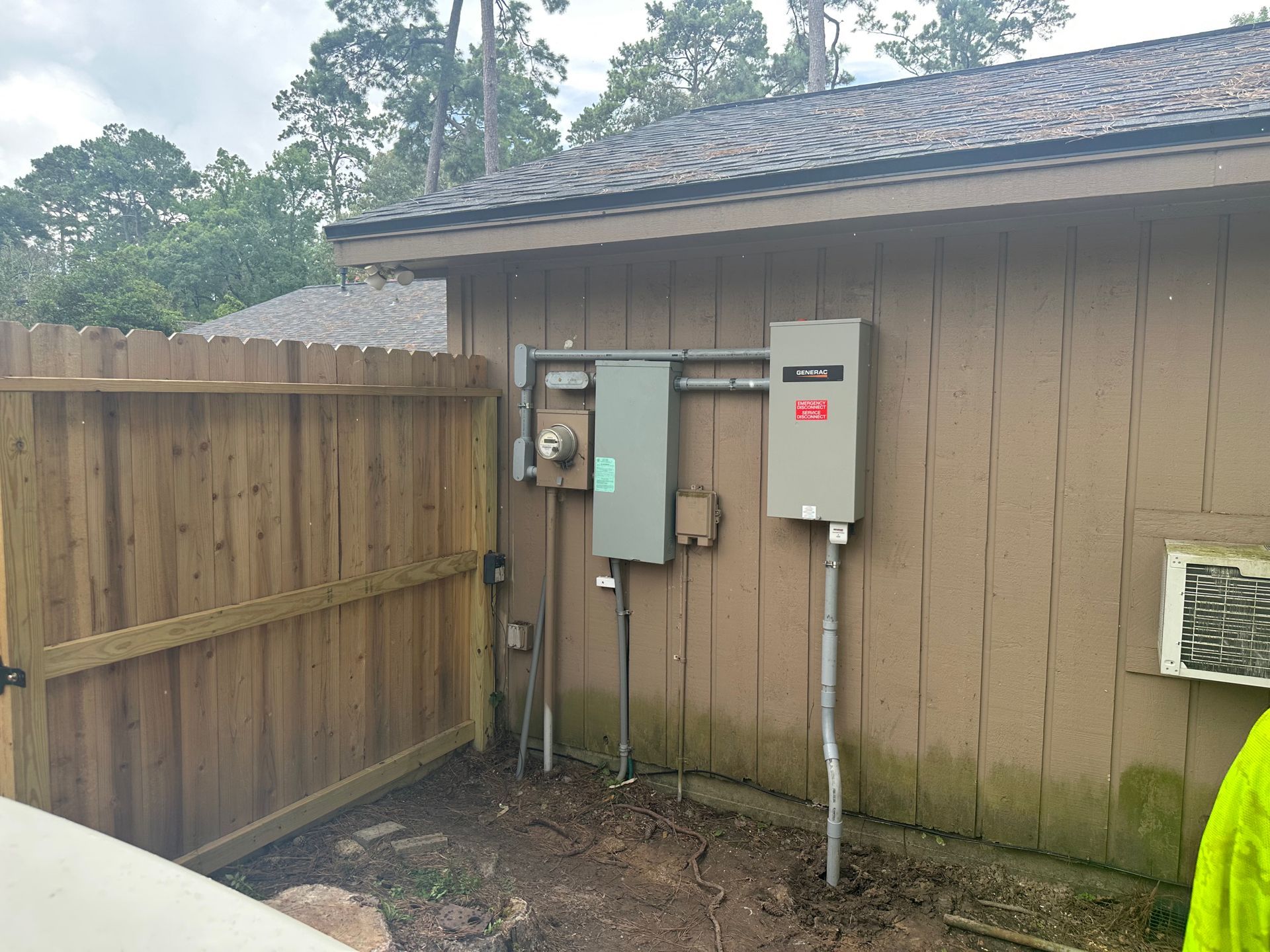 Electrical panel and meter on the side of a brown house next to a wooden fence.