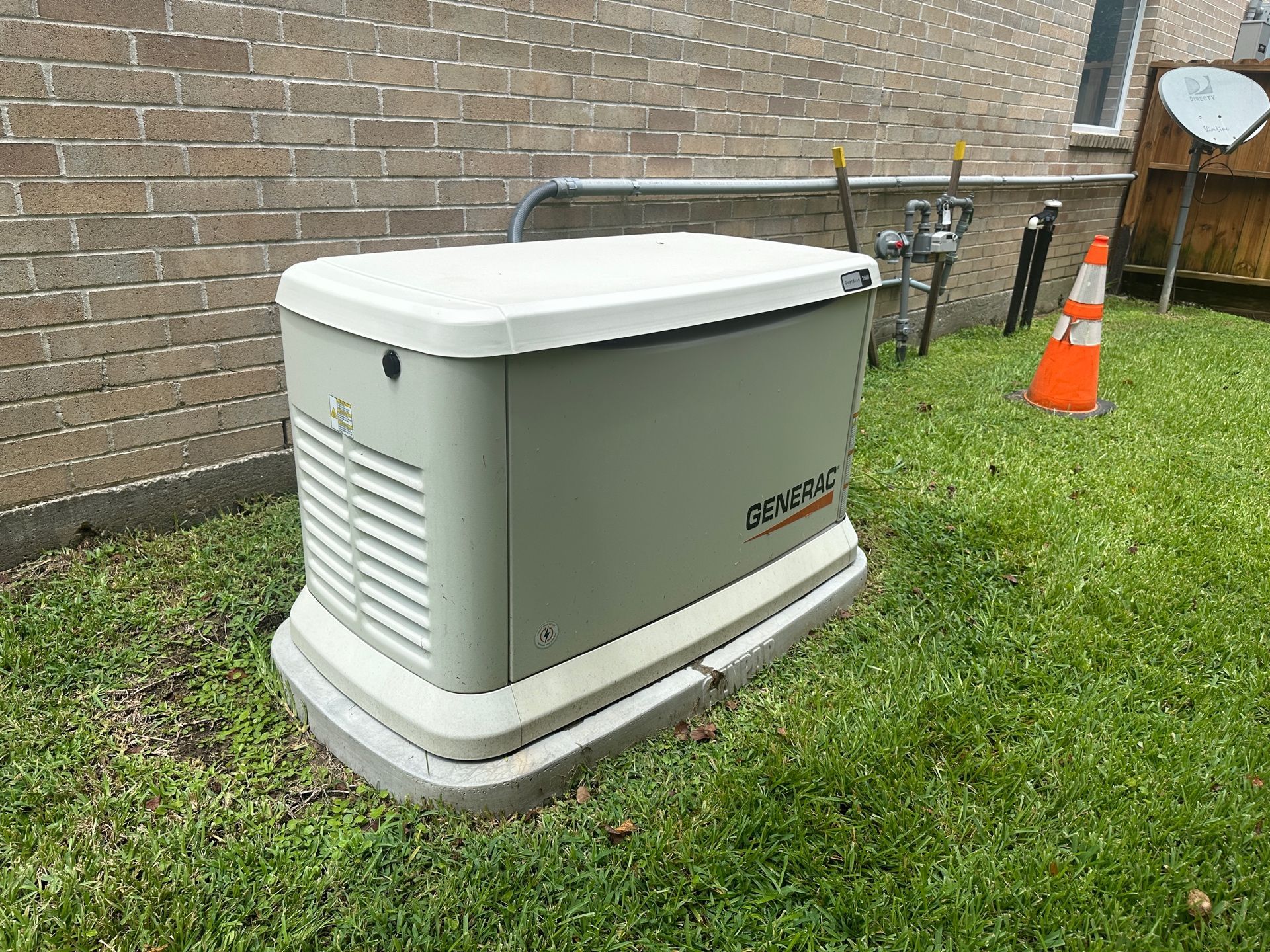 A Generac generator outside a brick building on a grassy lawn with a gas line and cone nearby.