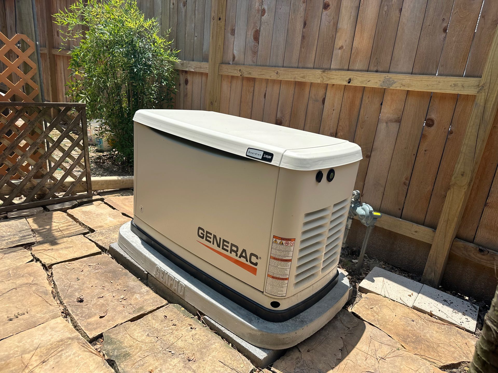 Generac home generator on a concrete pad, beige color. Located outdoors near a wooden fence and shrubbery.