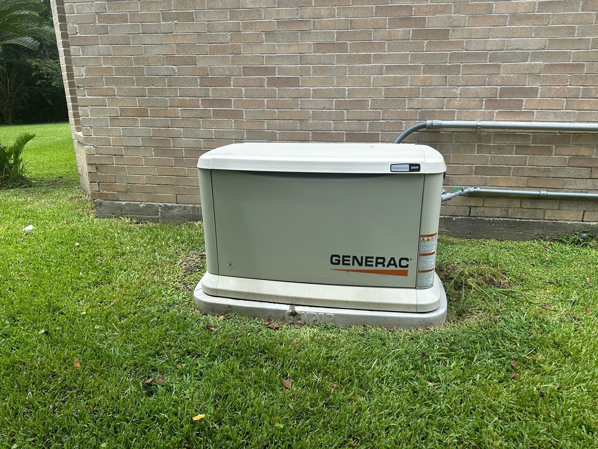 A Generac home generator is in green grass beside a brick wall, with metal conduit.