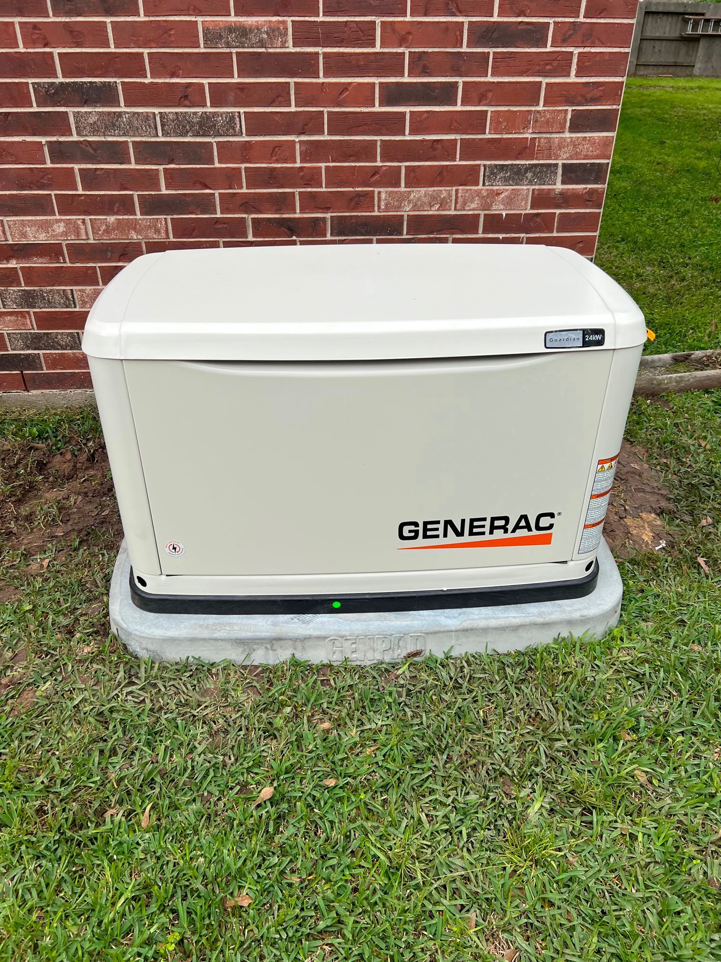 Generac home generator on a concrete base, next to a brick wall and grass.