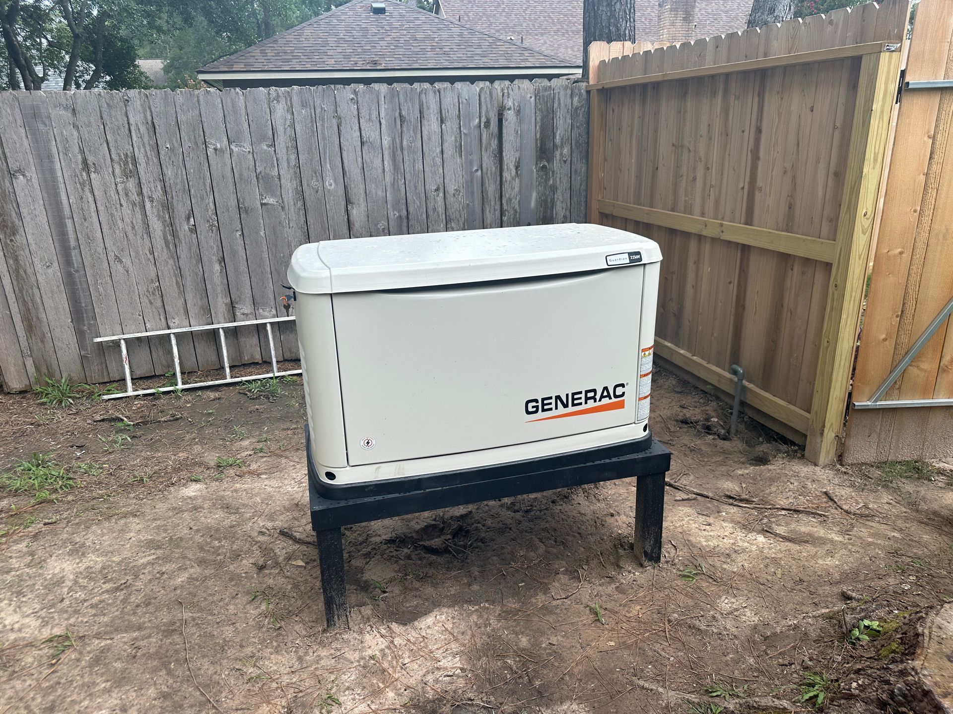 Generator on a black stand in a yard with a wooden fence. The generator is white with an orange logo.