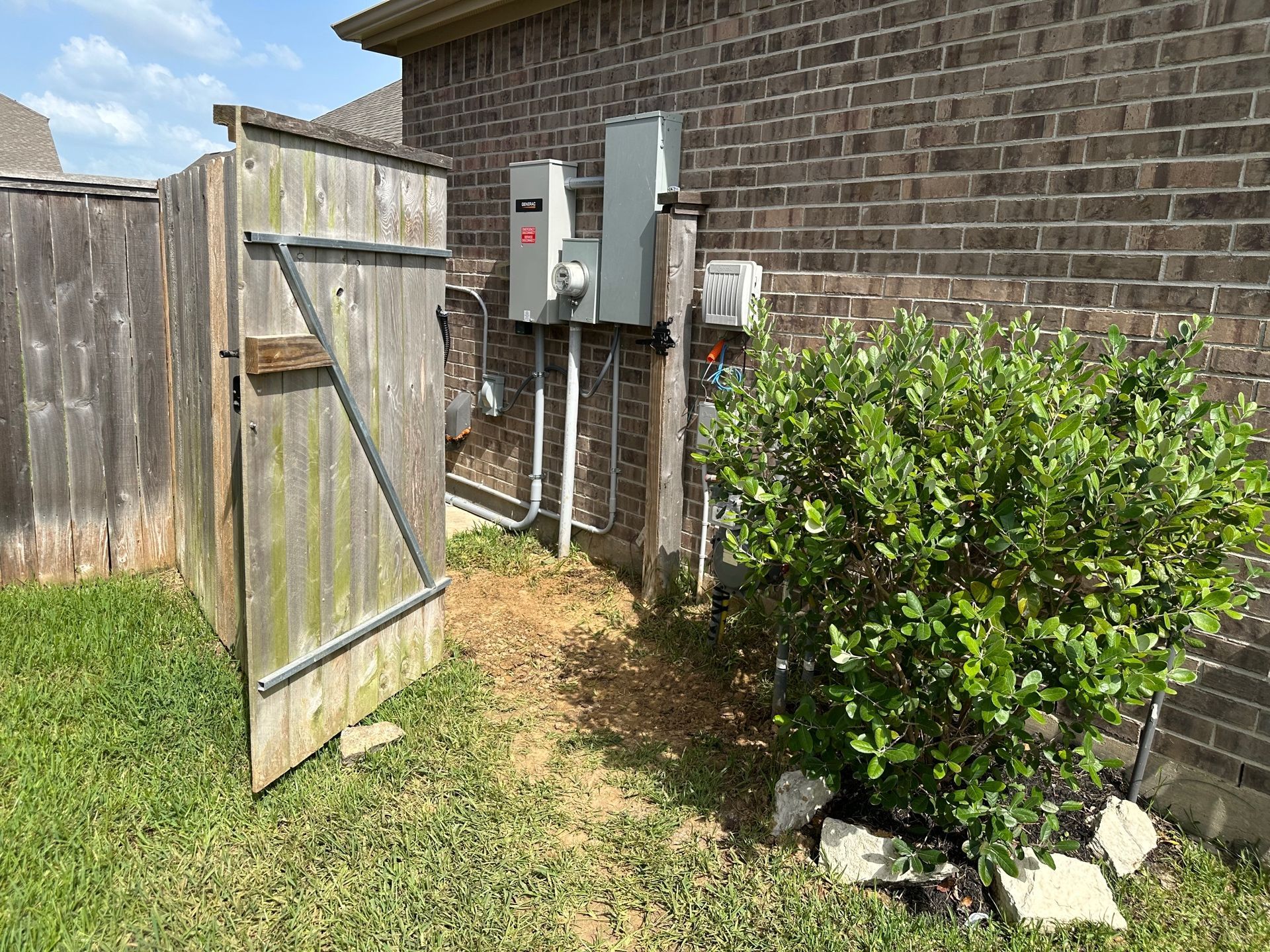 Wooden gate open to a backyard with electrical boxes on brick wall and a shrub.