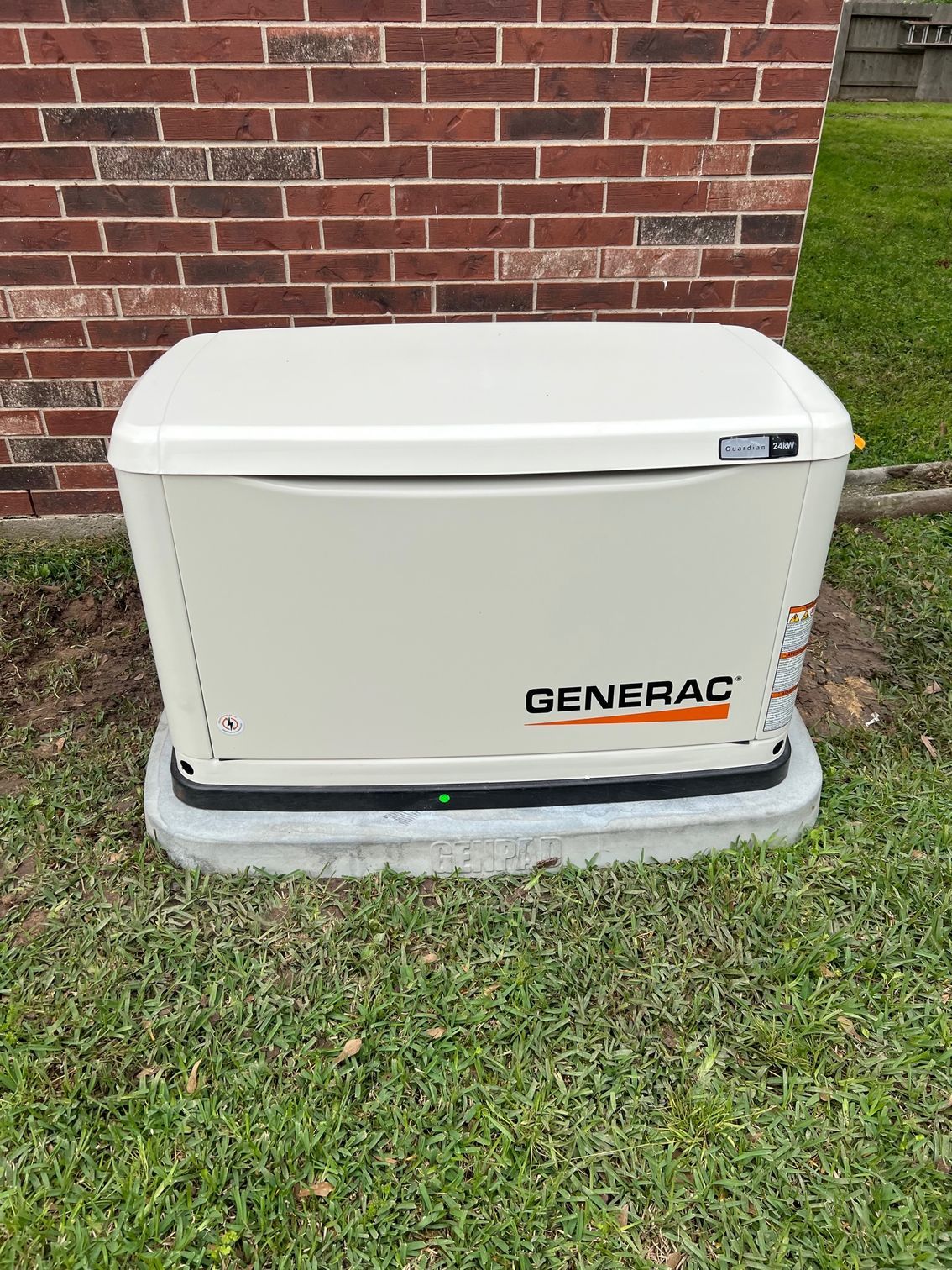 A Generac home standby generator on a concrete pad next to a brick wall and grass.