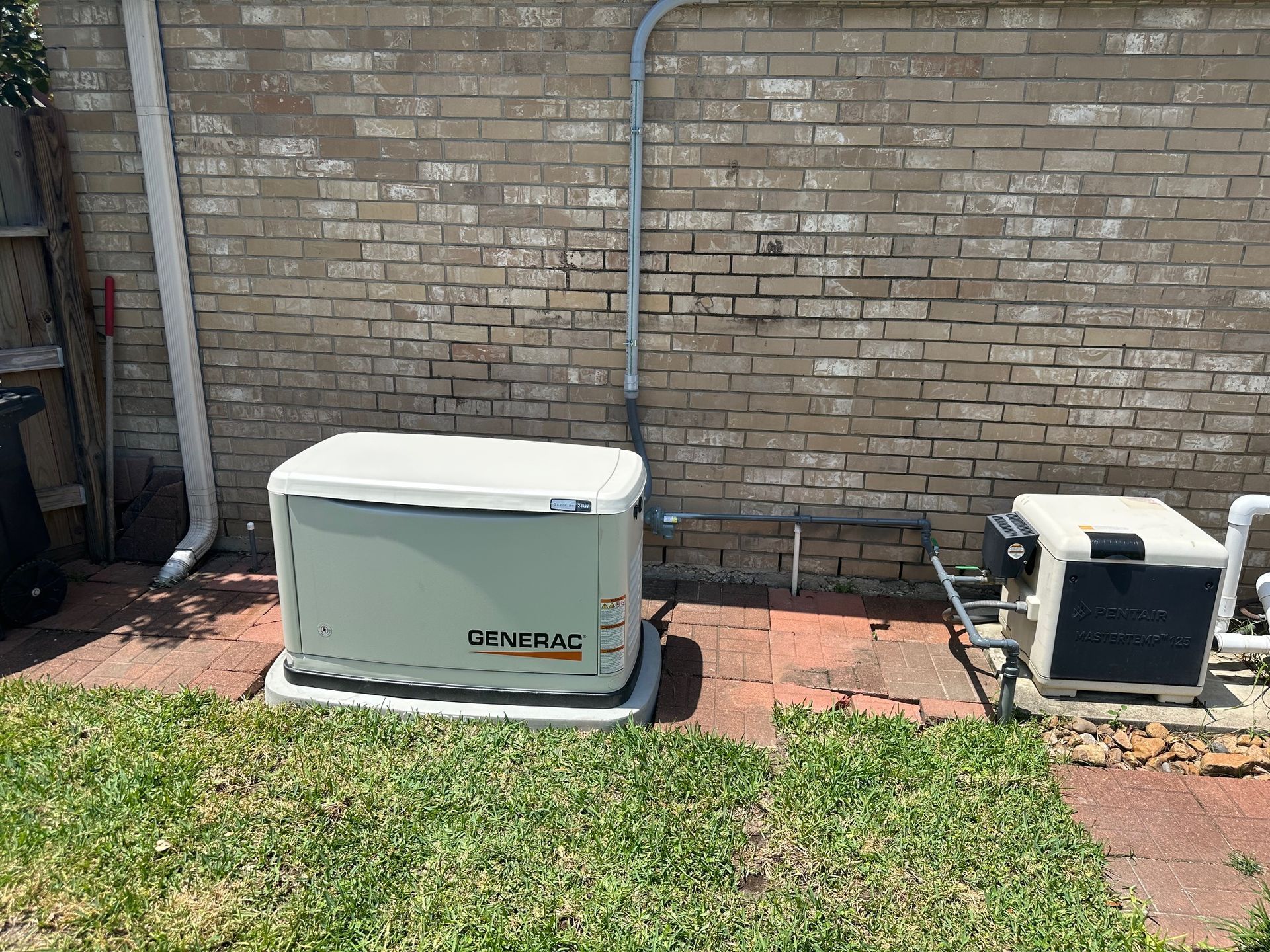 A Generac generator next to a brick wall and a pool pump, in a yard.
