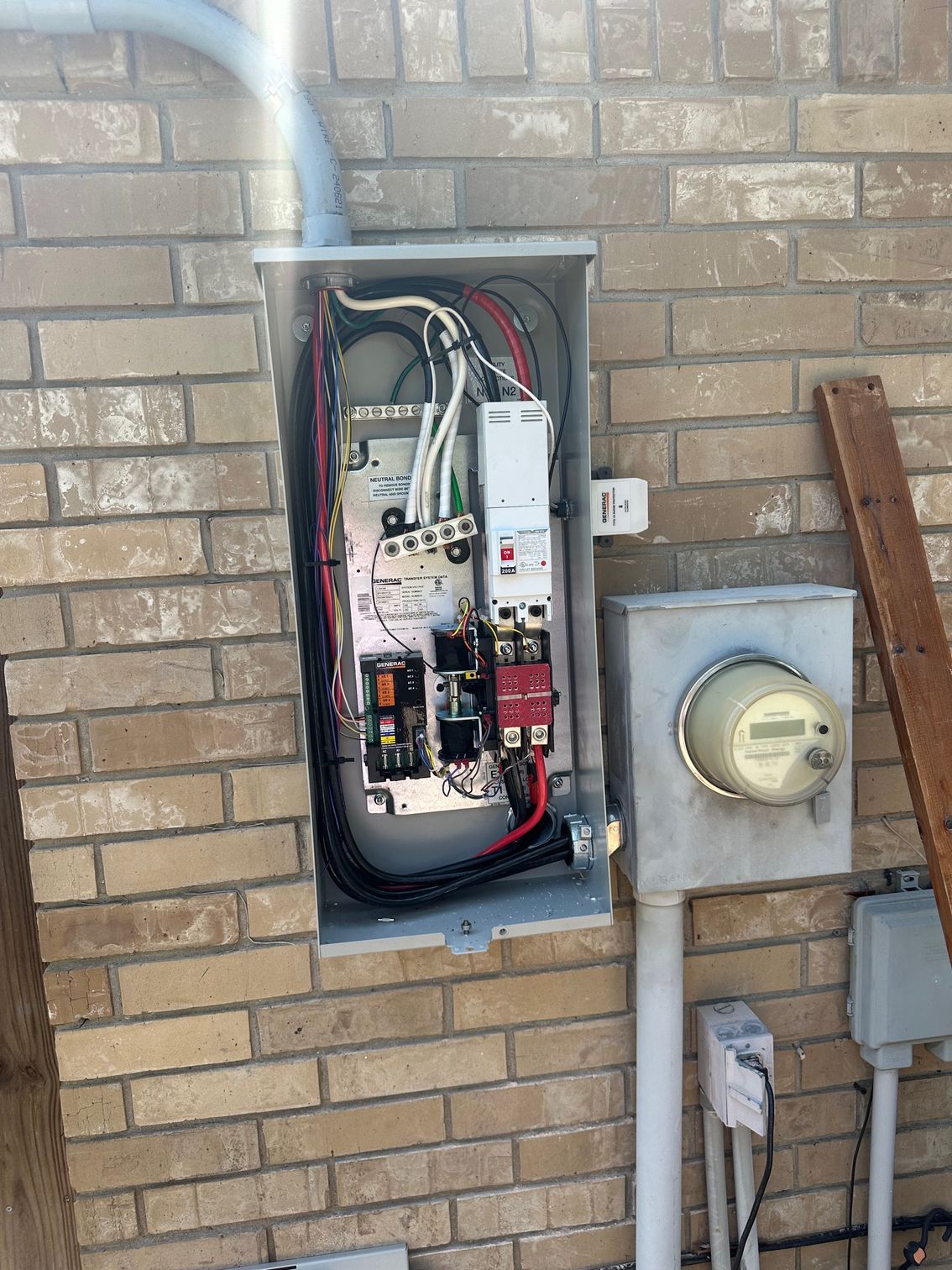Electrical panel mounted on brick wall, wires visible, next to a meter.