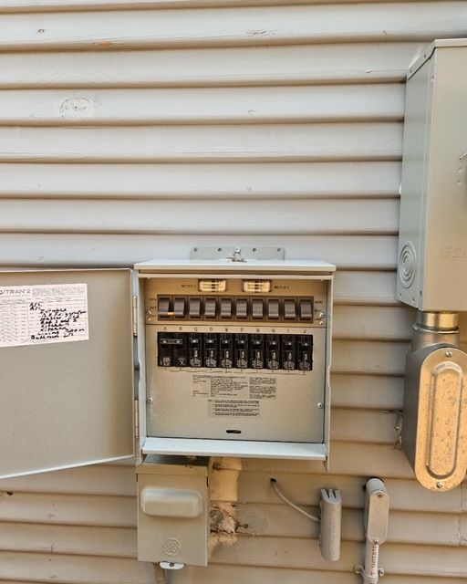 Exterior electrical panel mounted on beige siding. Open door reveals breakers; gray metal box, white pipes and cables nearby.