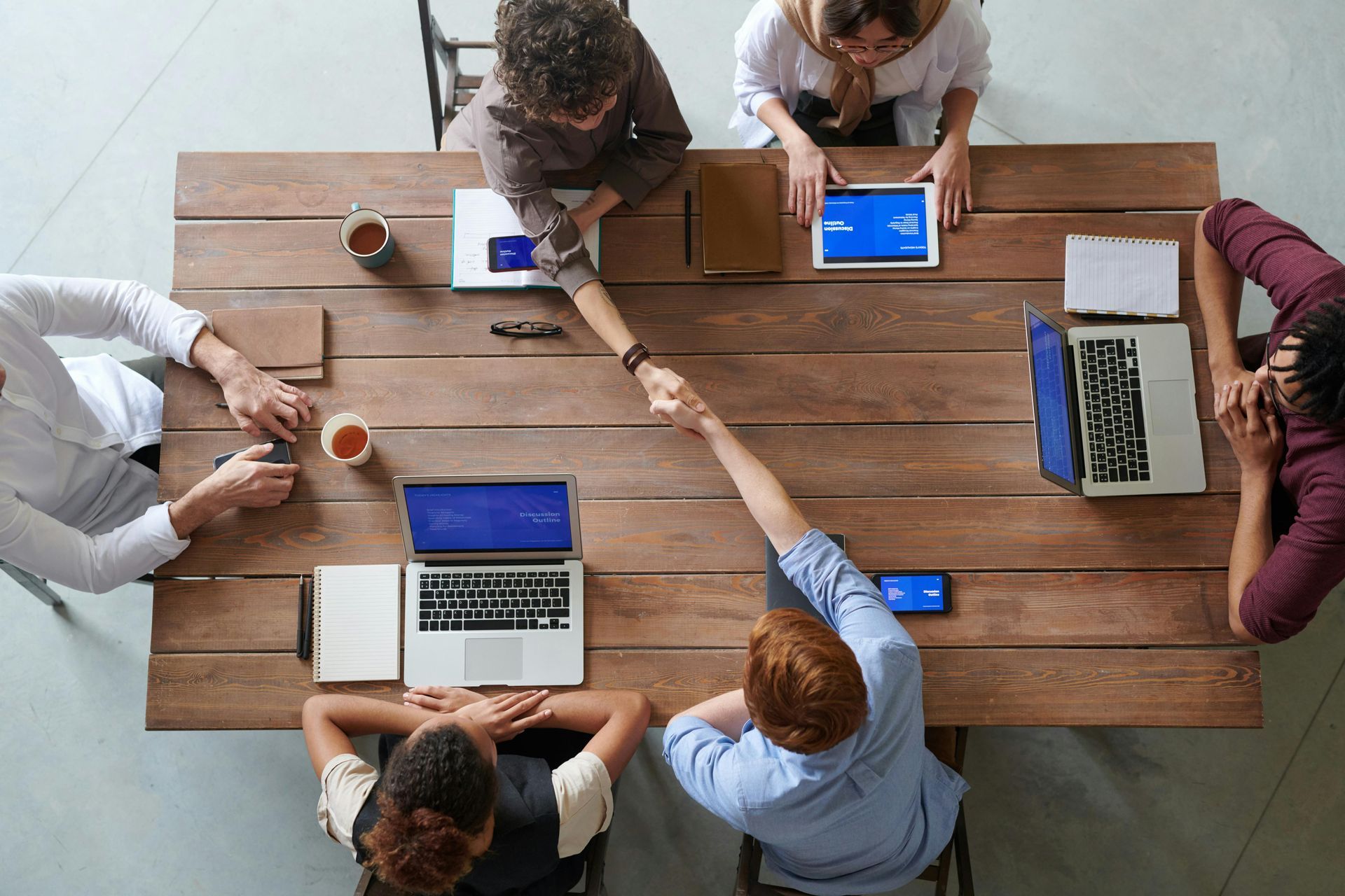 People shaking hands over a wooden table with laptops, tablets, and notebooks; office setting.