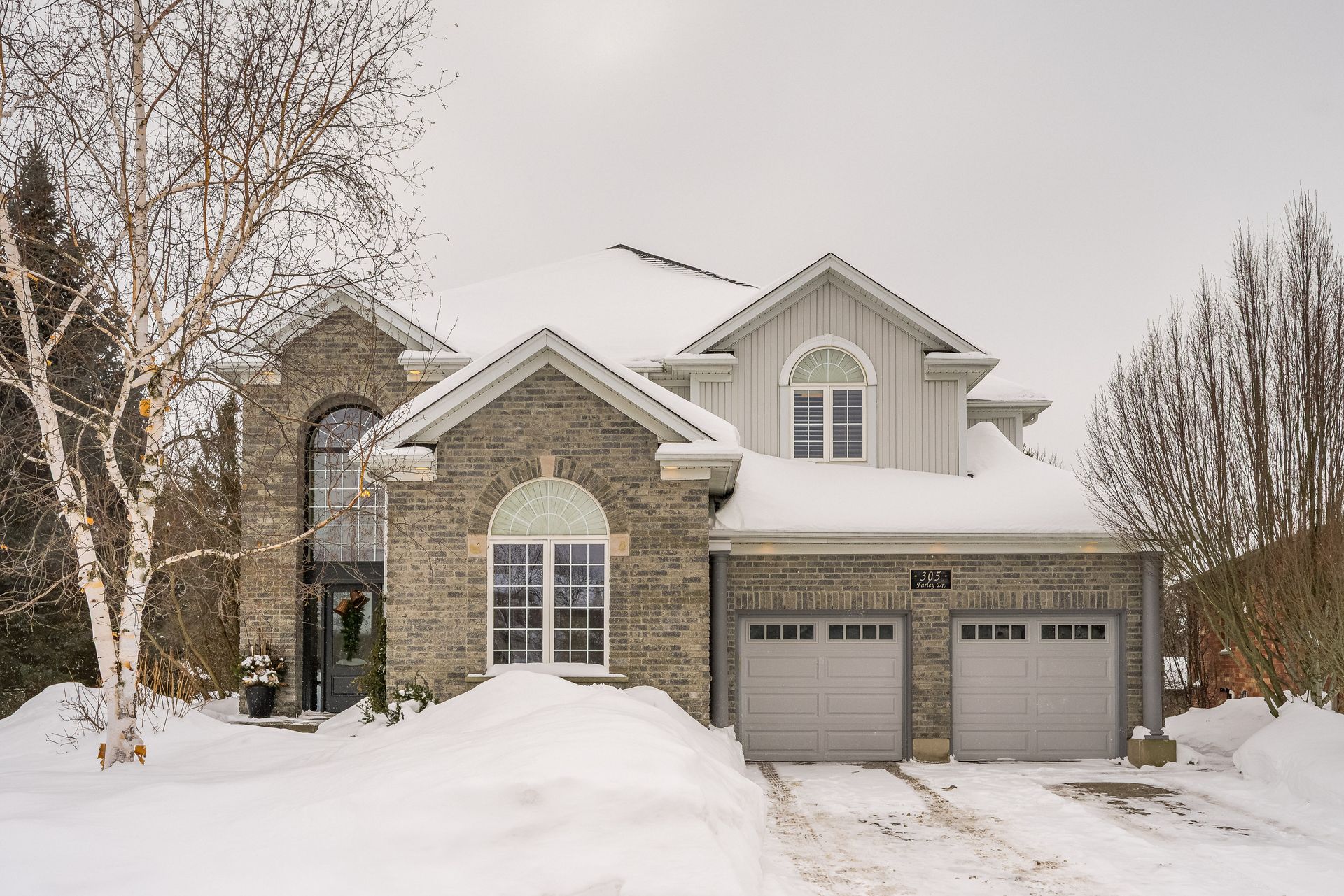 Snow-covered brick suburban house with two garage doors and bare trees in winter