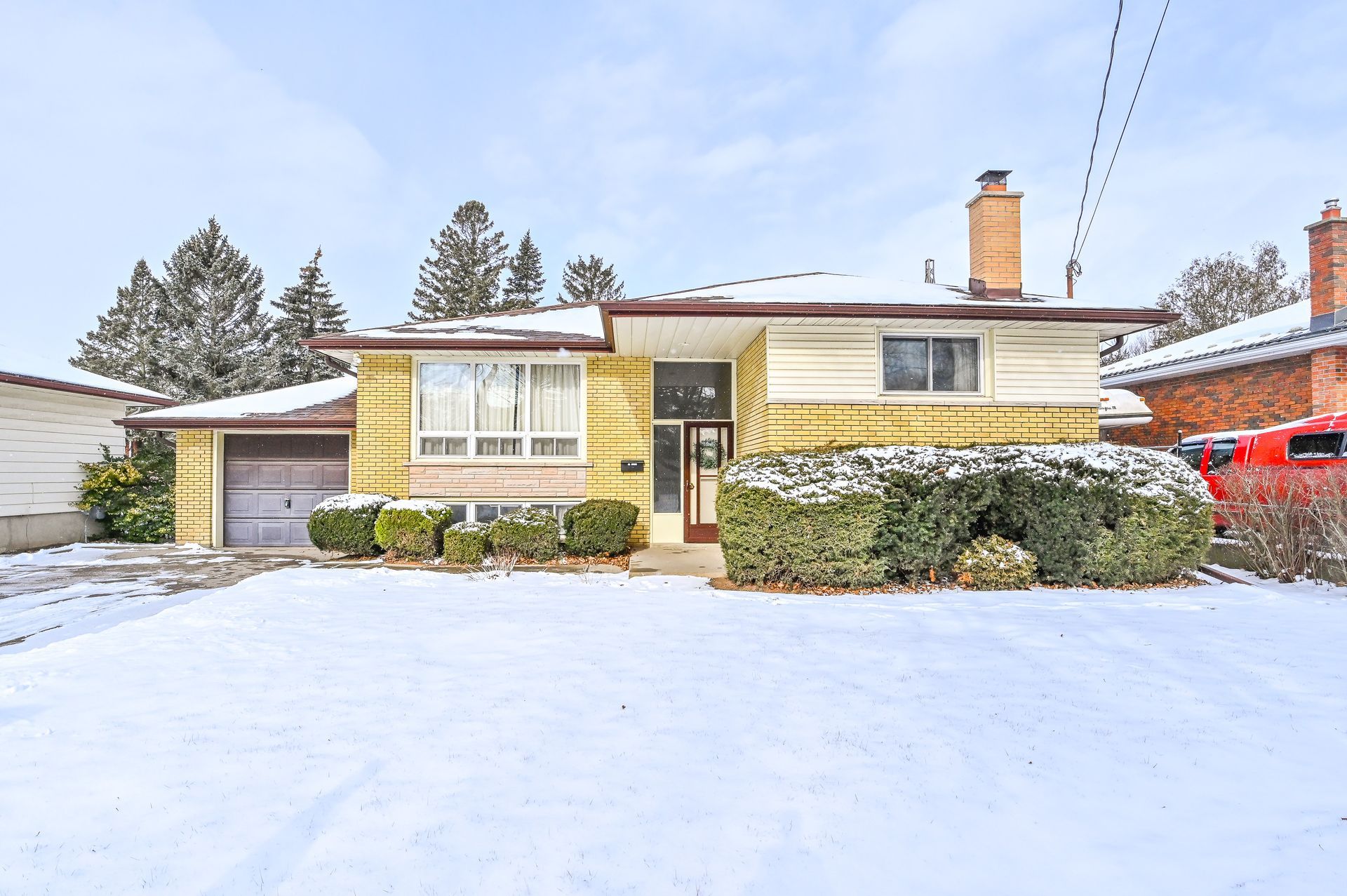 Snow-covered suburban house with yellow siding, a front walkway, and bushes under a cloudy winter sky