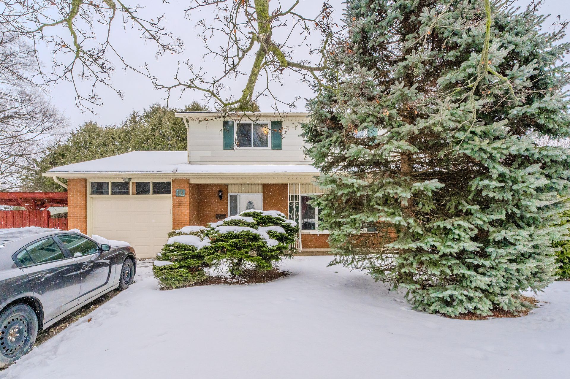 Snow-covered suburban house with a garage, large evergreen tree, and parked car in front