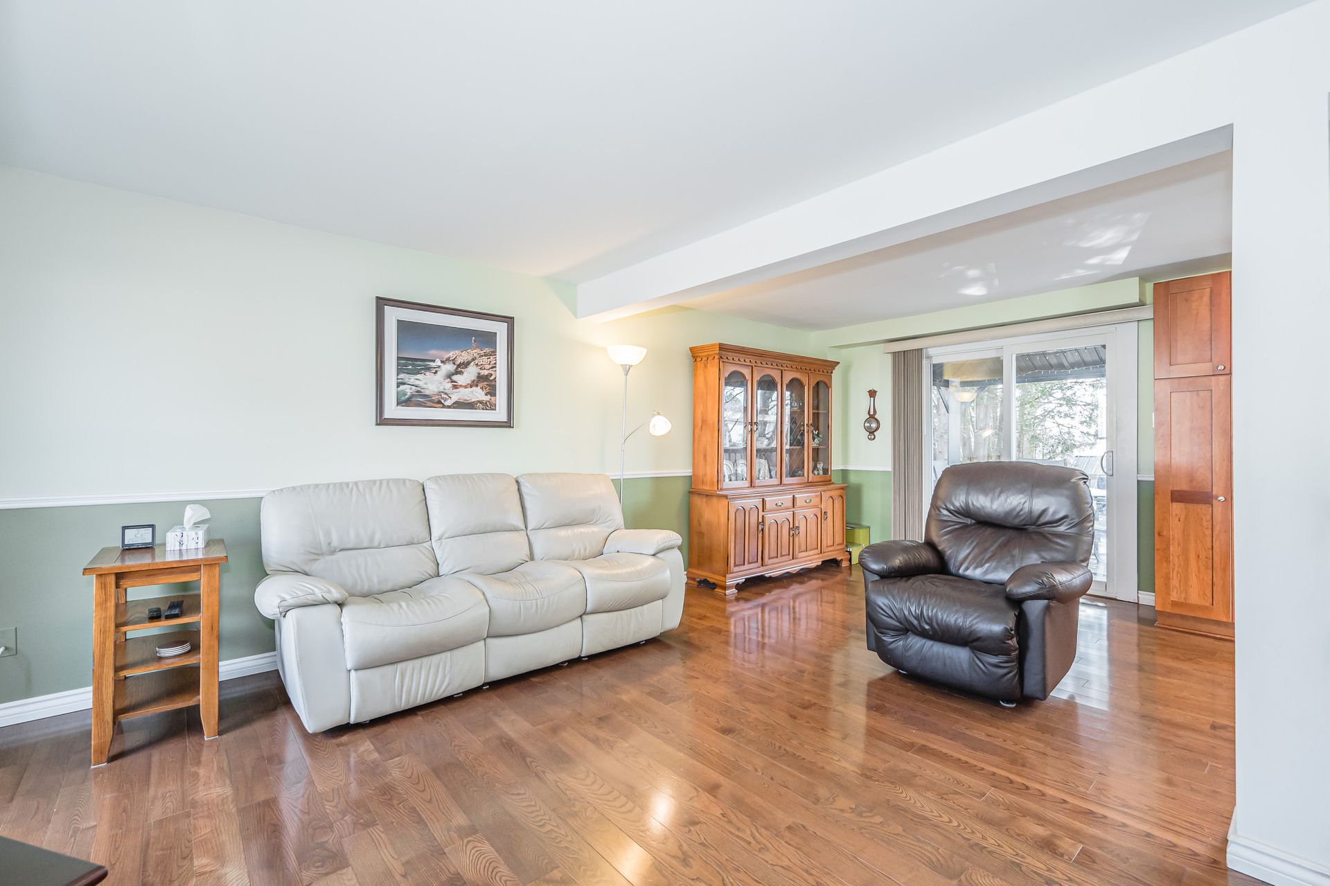 Living room with beige sofa, dark recliner, hardwood floors, and wood cabinets by a bright window