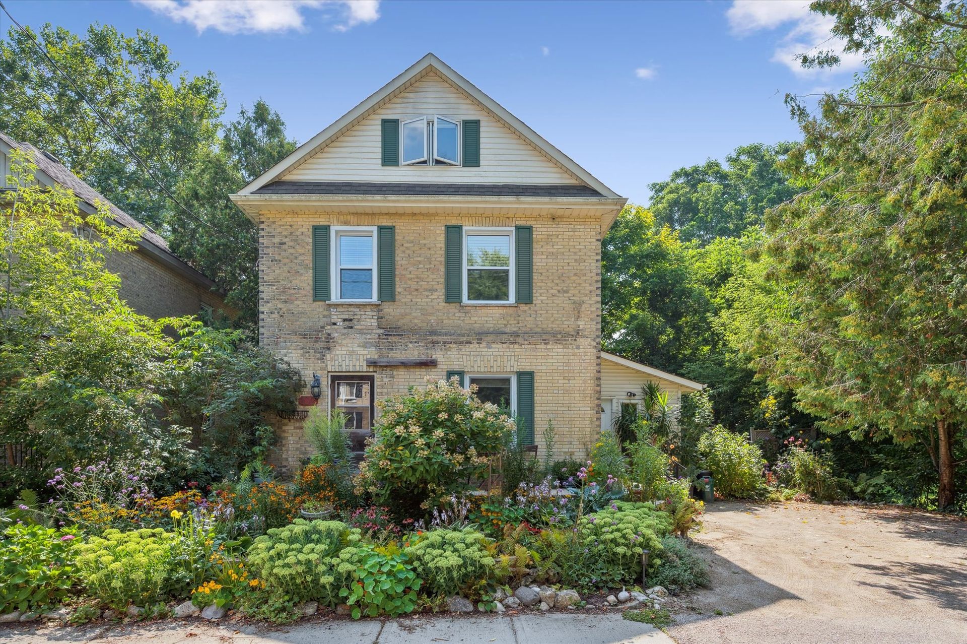 Two-story brick house with green shutters, surrounded by lush garden and trees under a blue sky