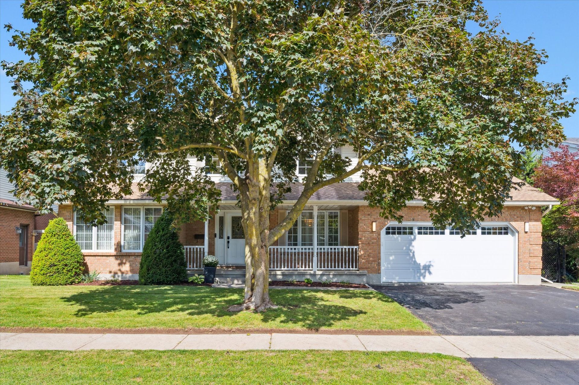 Suburban brick house with a large front tree, lawn, and white garage door under a clear sky