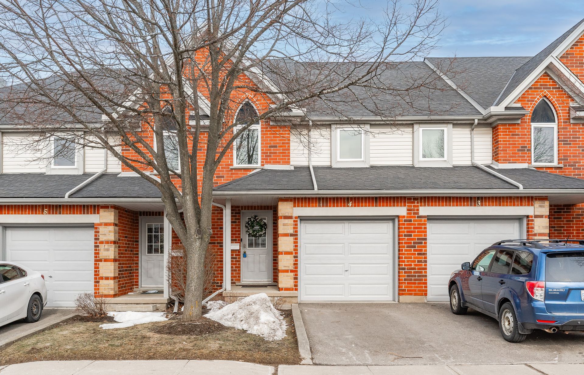 Row of brick townhouses with attached garages, a bare tree in front, and cars parked outside in winter.