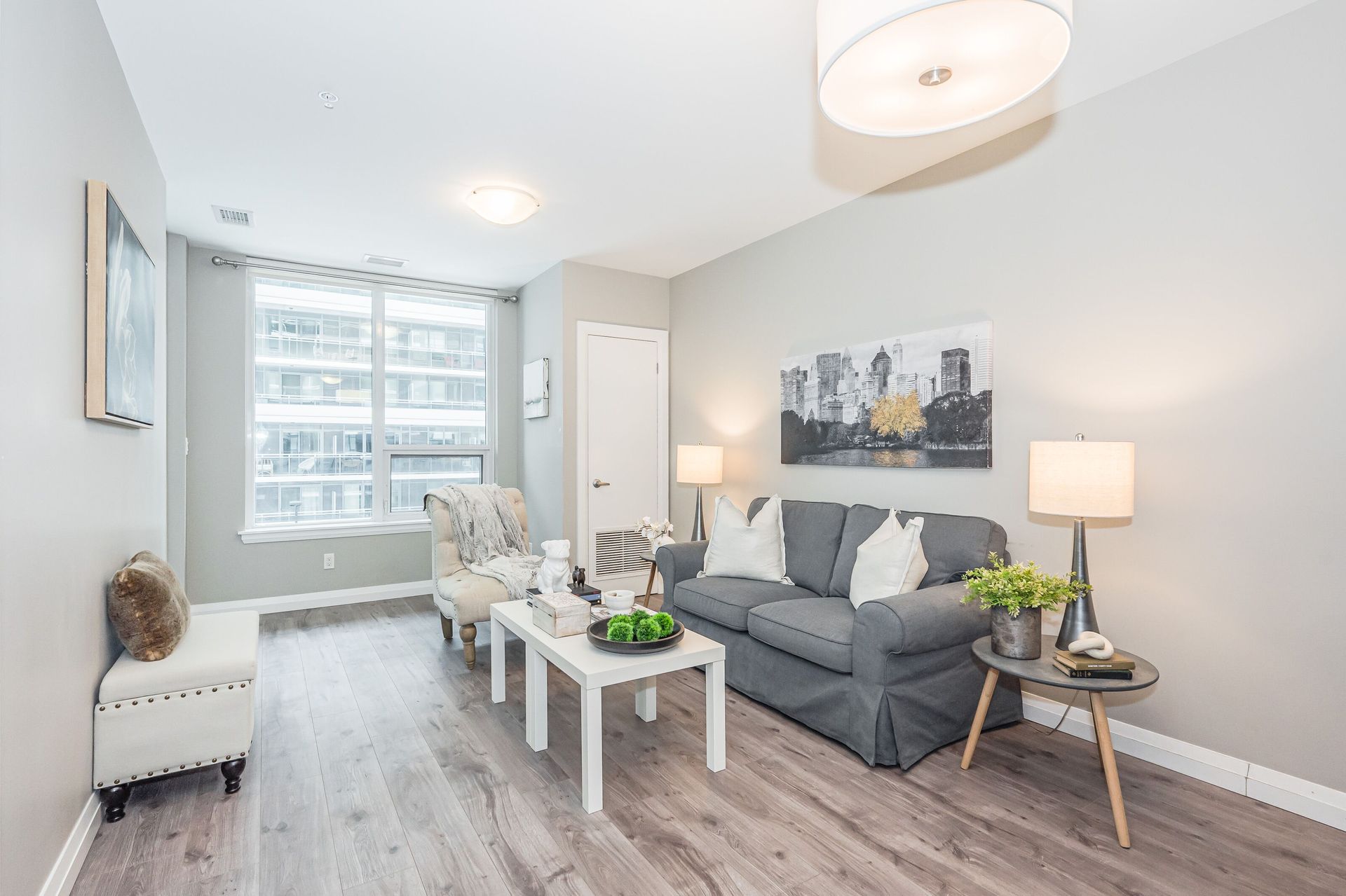Bright living room with gray sofa, white coffee table, armchair, lamp, and large window over wood floors.