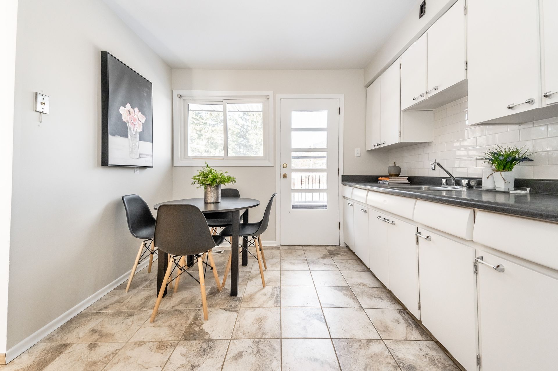 Bright kitchen with white cabinets, marble floor, black table and chairs, and a glass door at the end