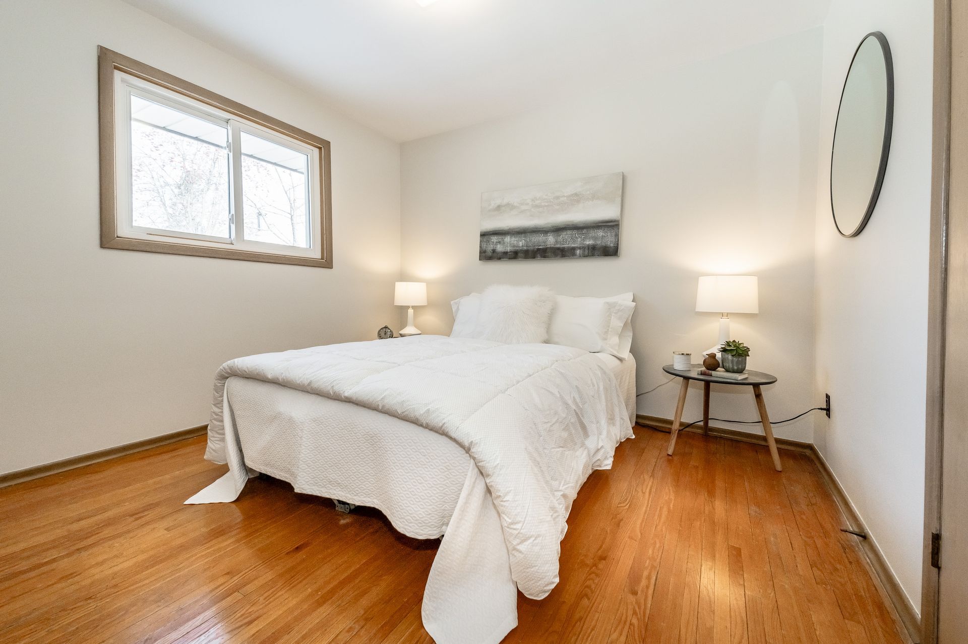 Bright bedroom with a white bed, wooden floor, bedside lamps, and a window on the left.