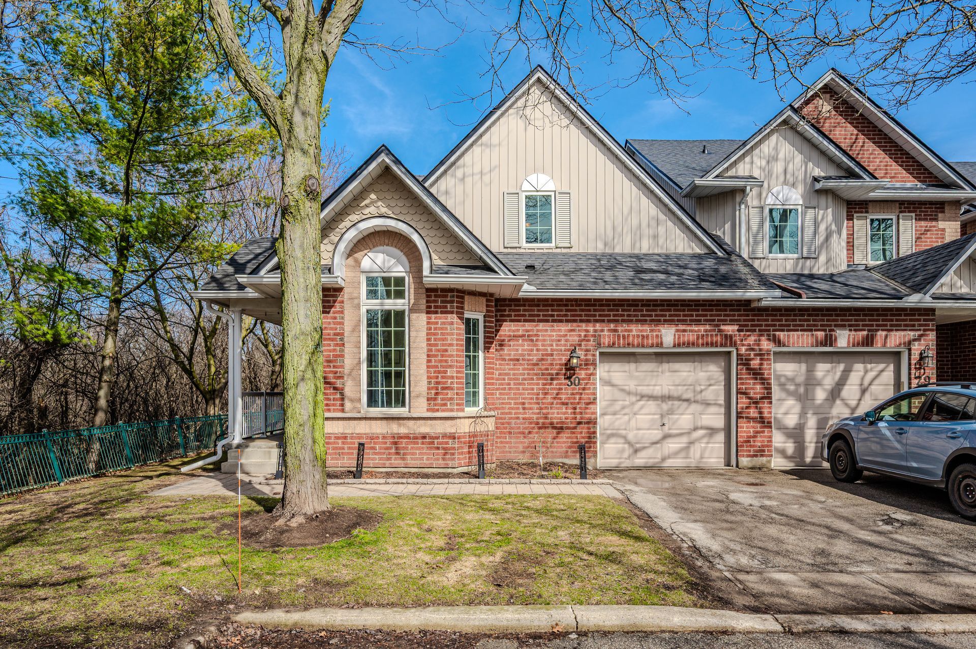 Suburban brick townhouse with attached garage, driveway, and a car parked outside under a bright sky