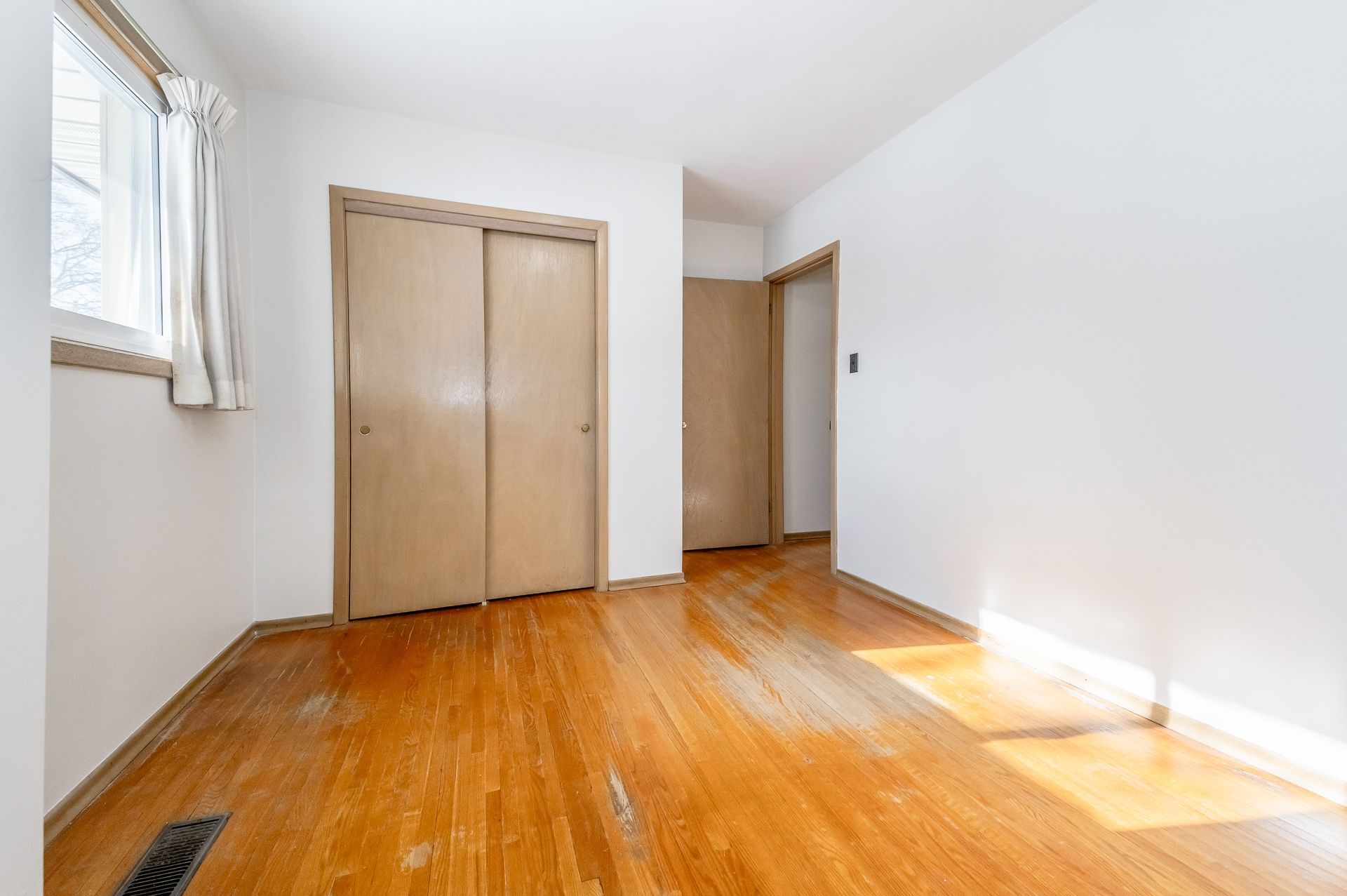Empty sunlit bedroom with hardwood floor, white walls, a window, and a wooden closet door