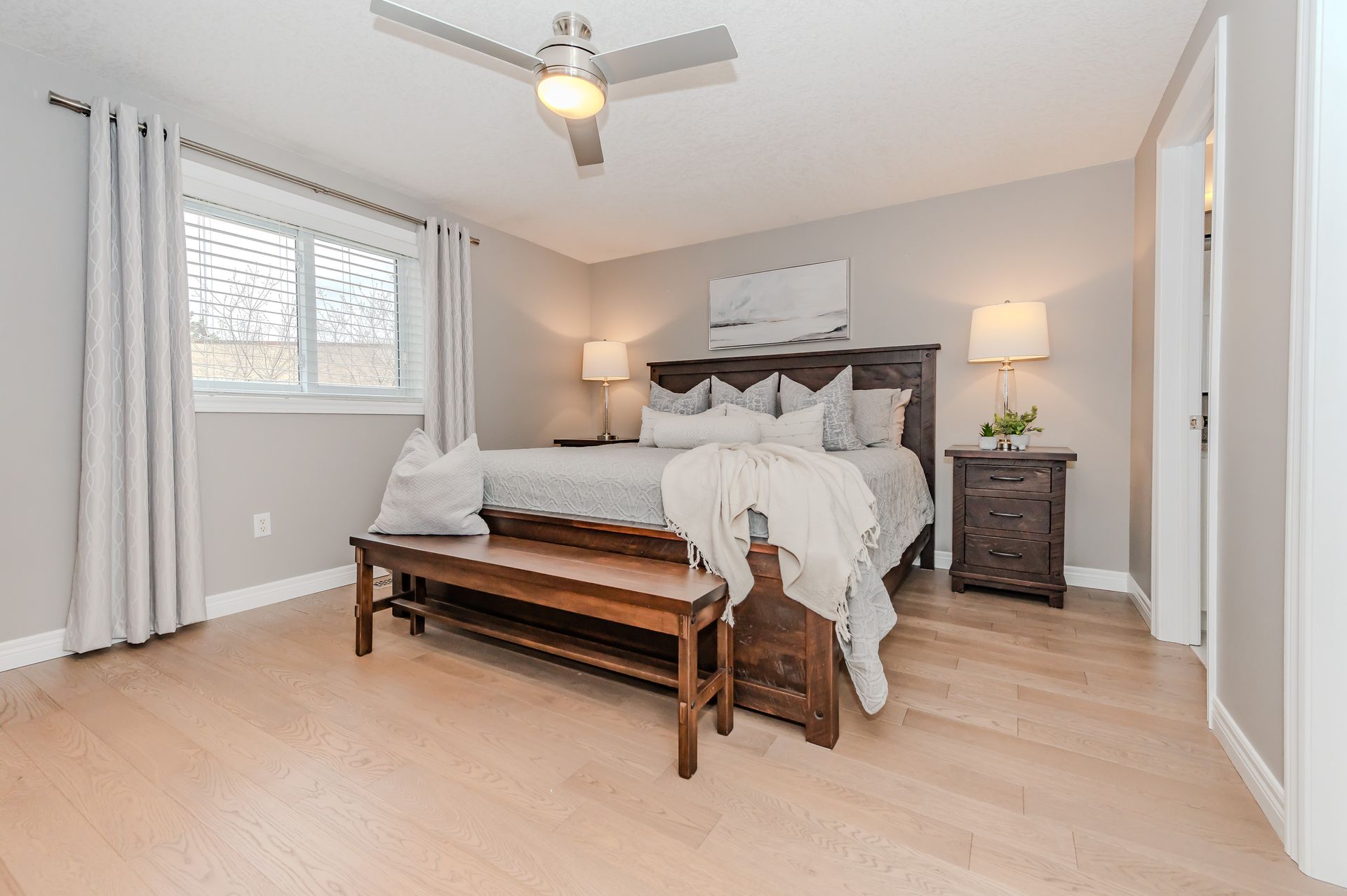 Bright bedroom with a wooden bed, bench, bedside lamps, and light curtains on a pale floor