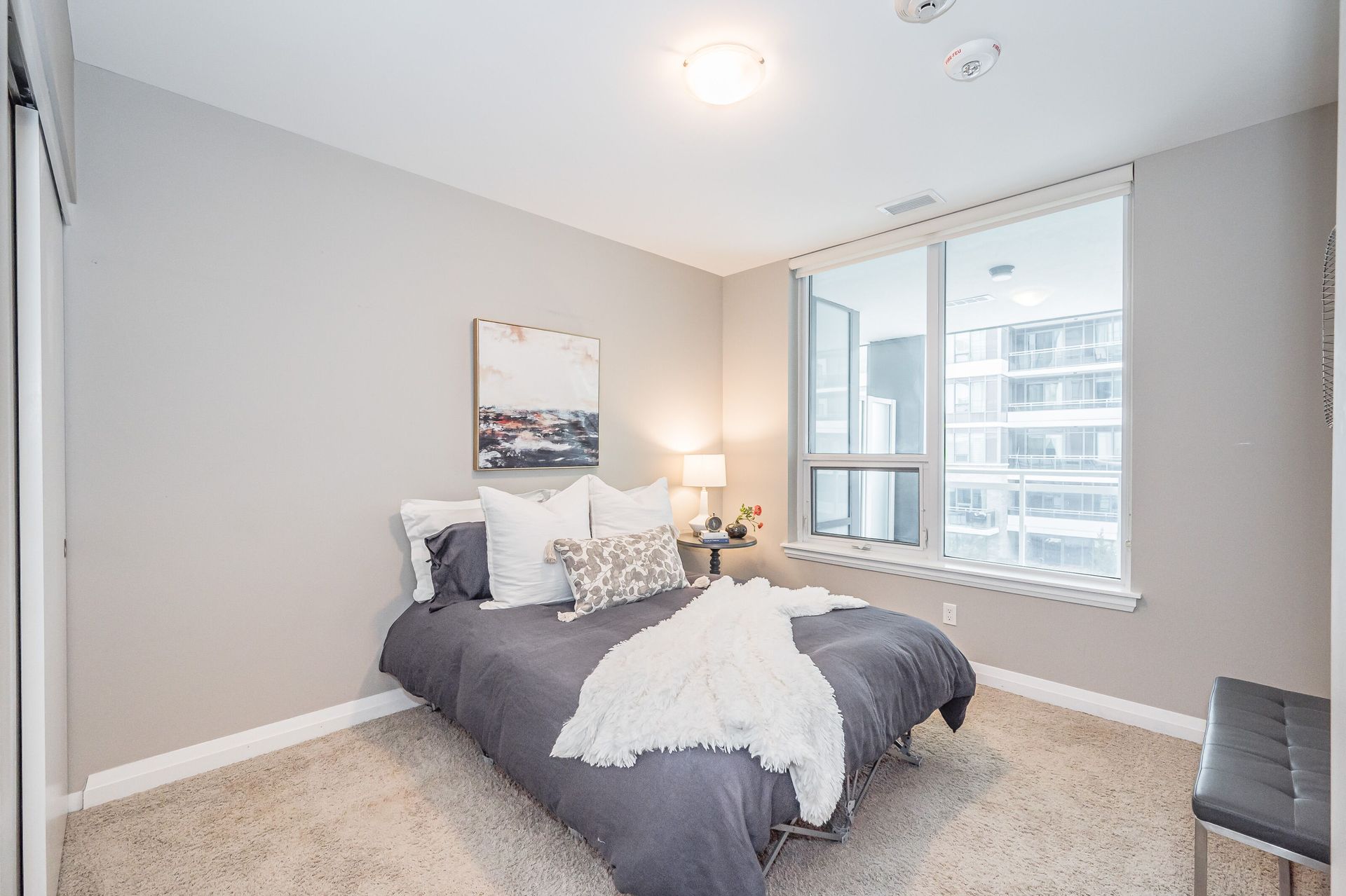 Bright bedroom with a gray bed, white decor, and a large window overlooking city buildings