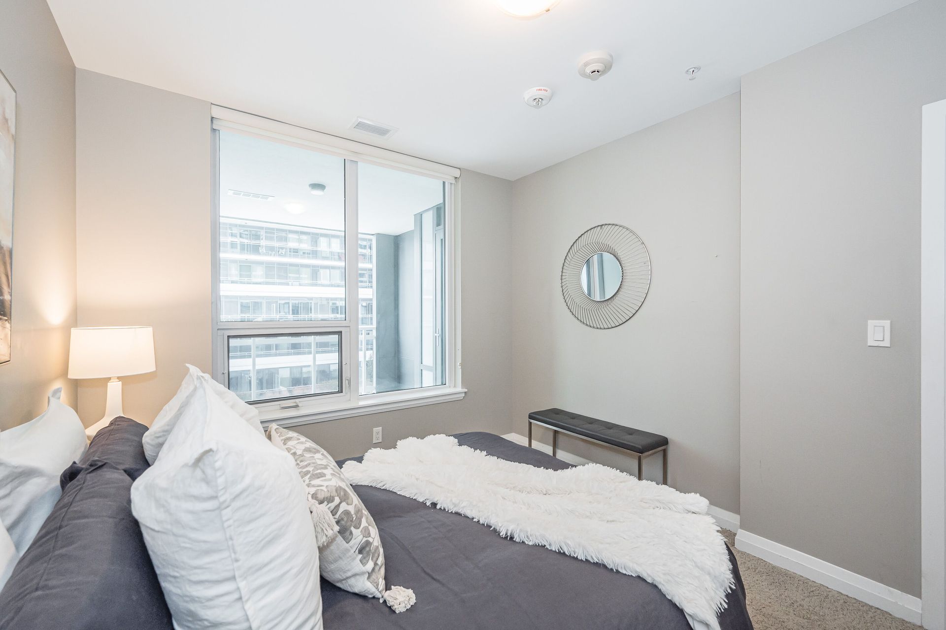 Bright bedroom with gray walls, white bedding, a window seat, and a round wall mirror