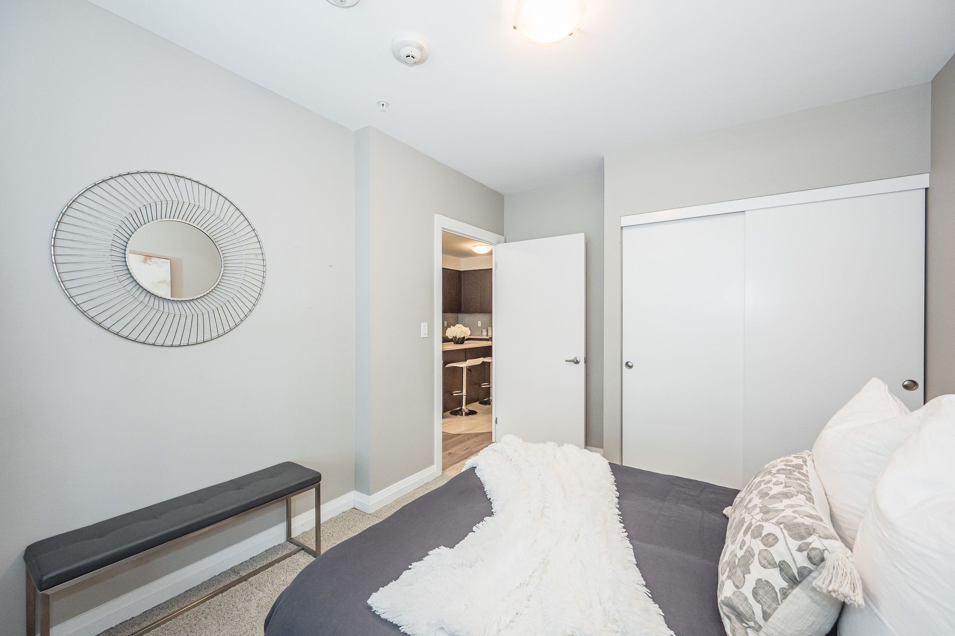 Bright bedroom with gray walls, white closet doors, bench, wall mirror, and bed with white bedding
