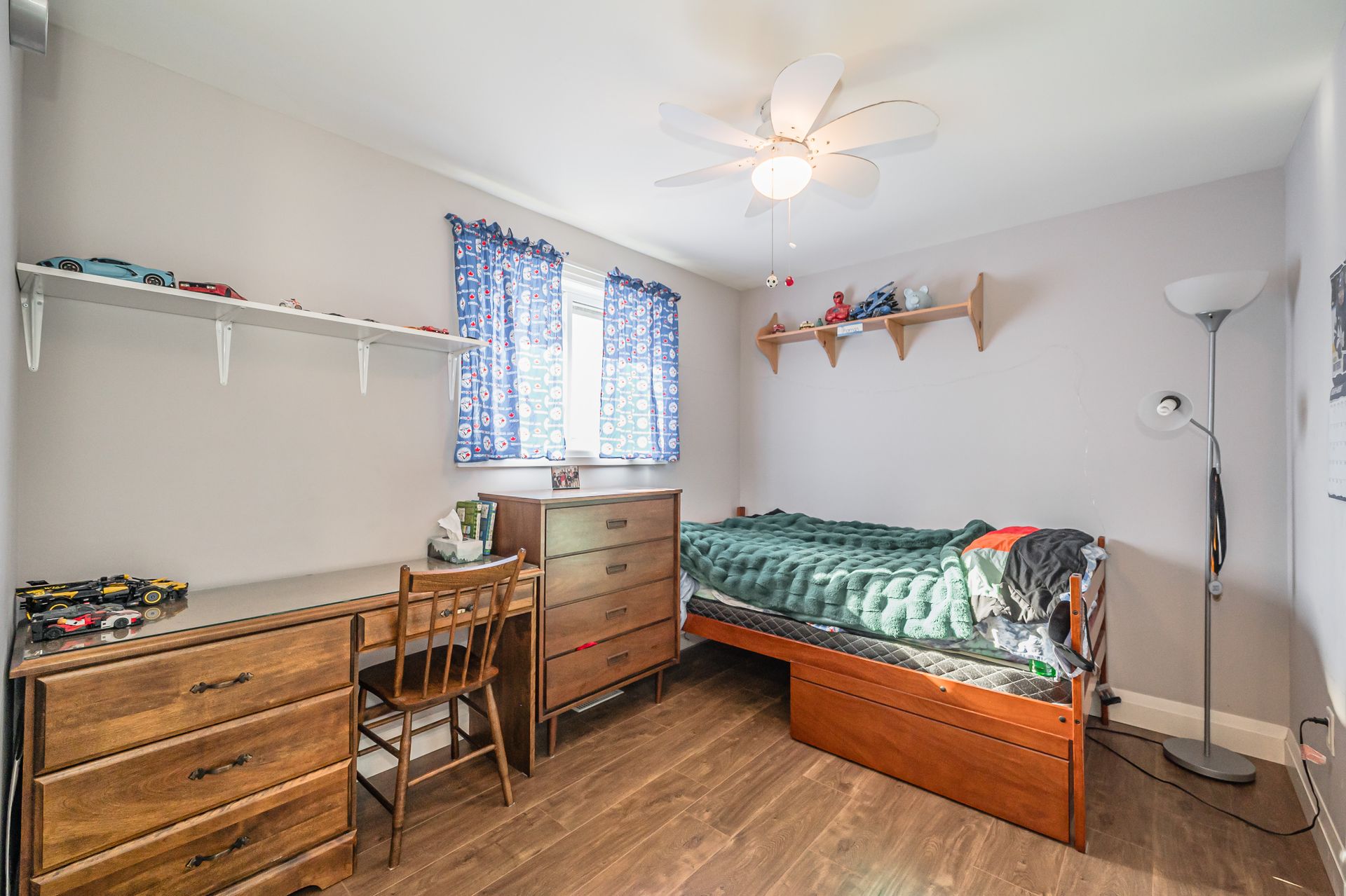 Small bedroom with twin bed, dresser, desk, shelves, and blue curtains in a bright white room