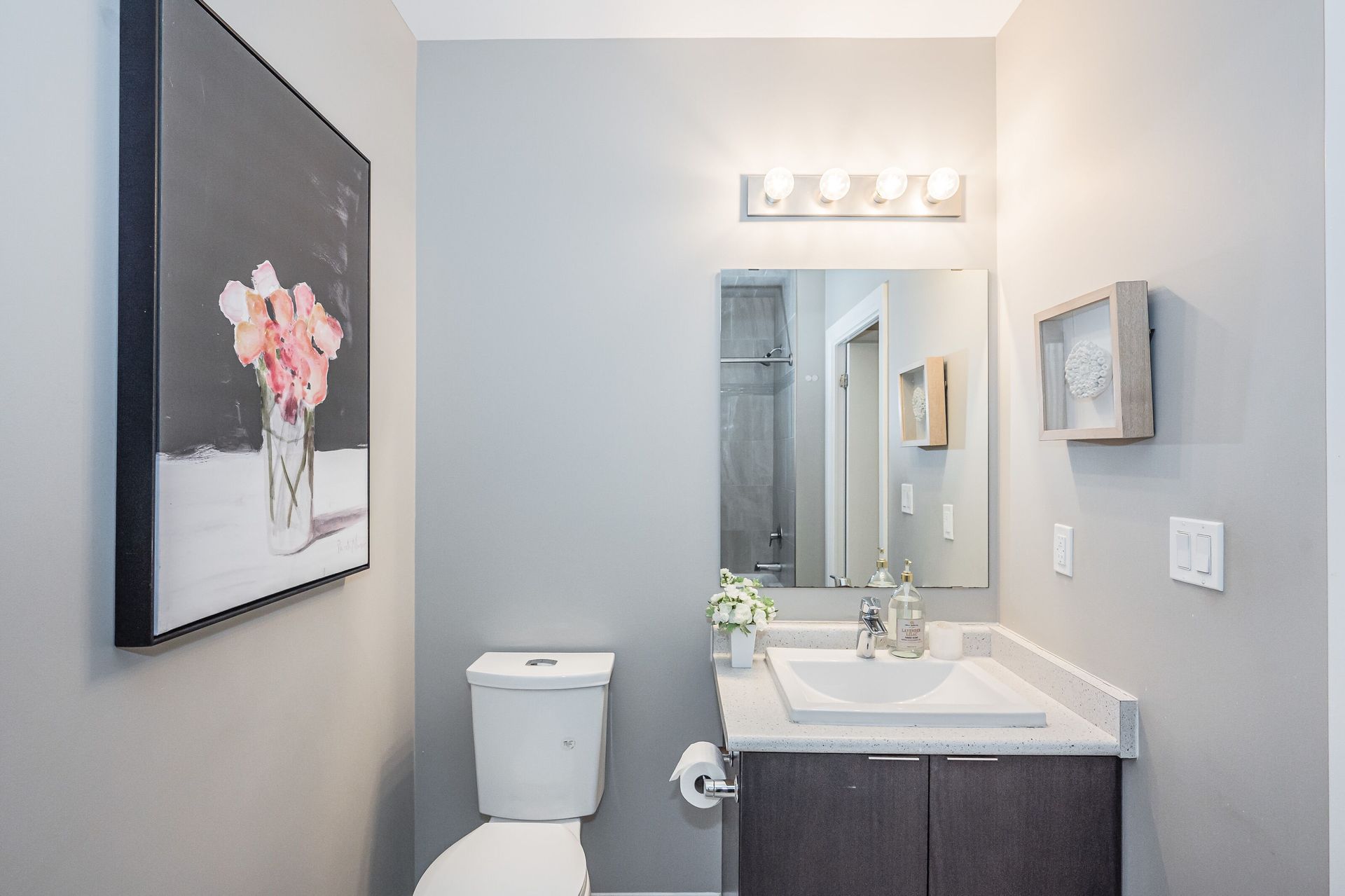 Small bathroom with toilet, vanity sink, mirror, wall art, and light gray walls.