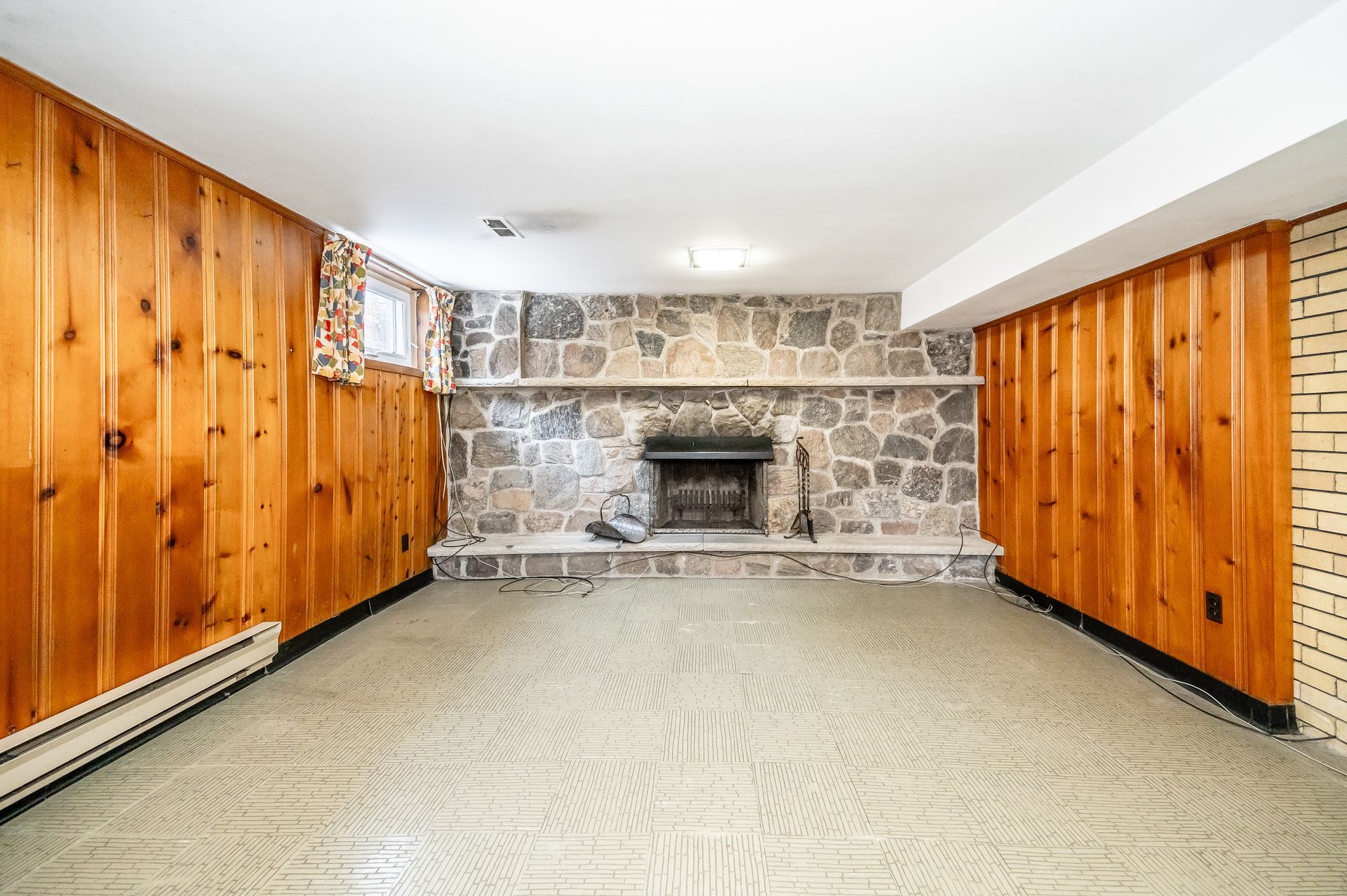 Empty basement rec room with wood-paneled walls and a stone fireplace at the far end