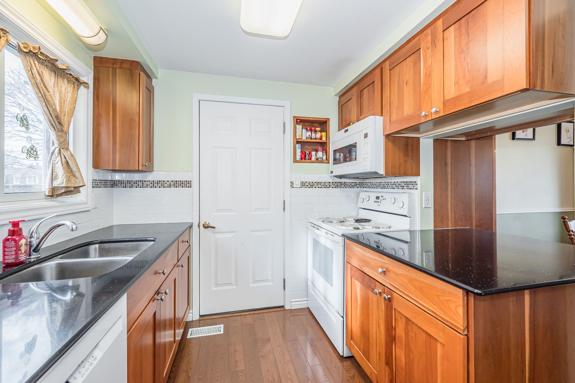 Compact kitchen with wood cabinets, black counters, white appliances, and a tiled backsplash.