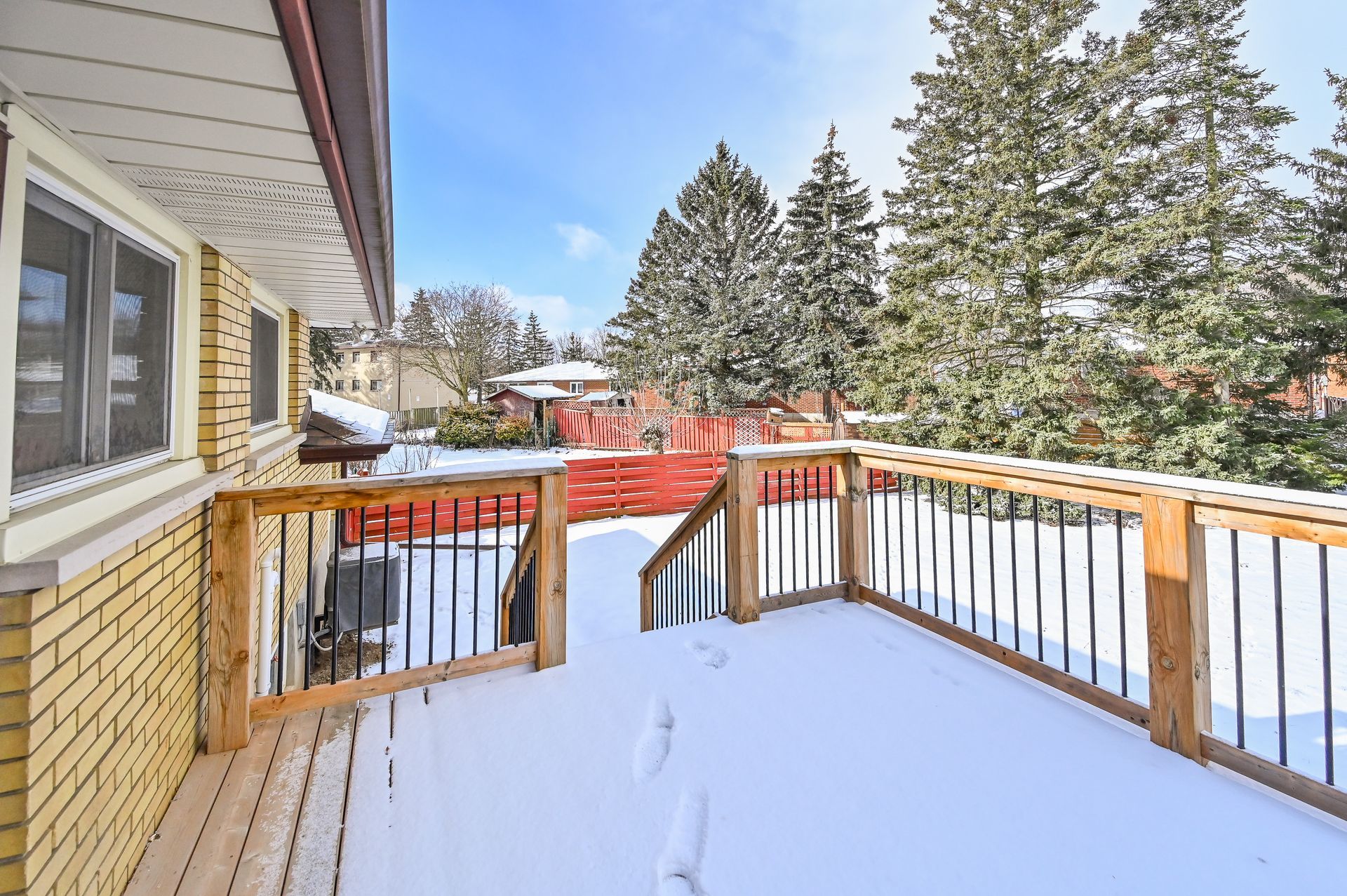 Snow-covered wooden deck beside a house, with railing and evergreen trees in the background.