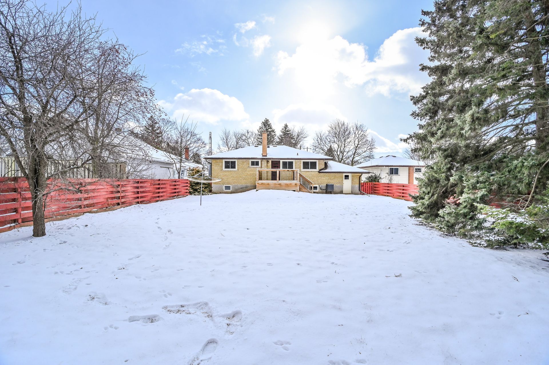 Snow-covered backyard with a yellow house, red fence, and trees under a bright winter sky