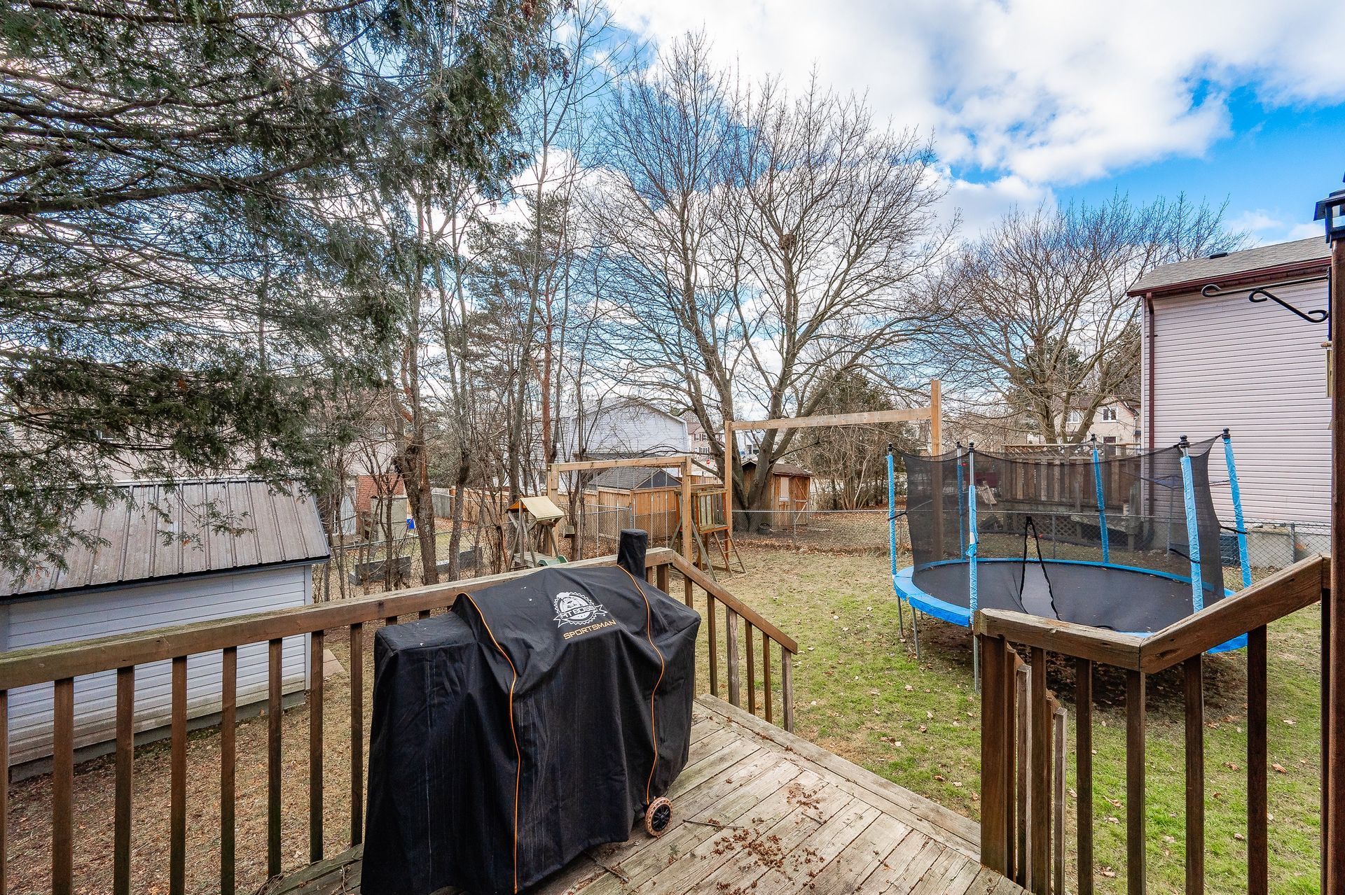 Backyard deck with covered grill, trampoline, leafless trees, and cloudy sky