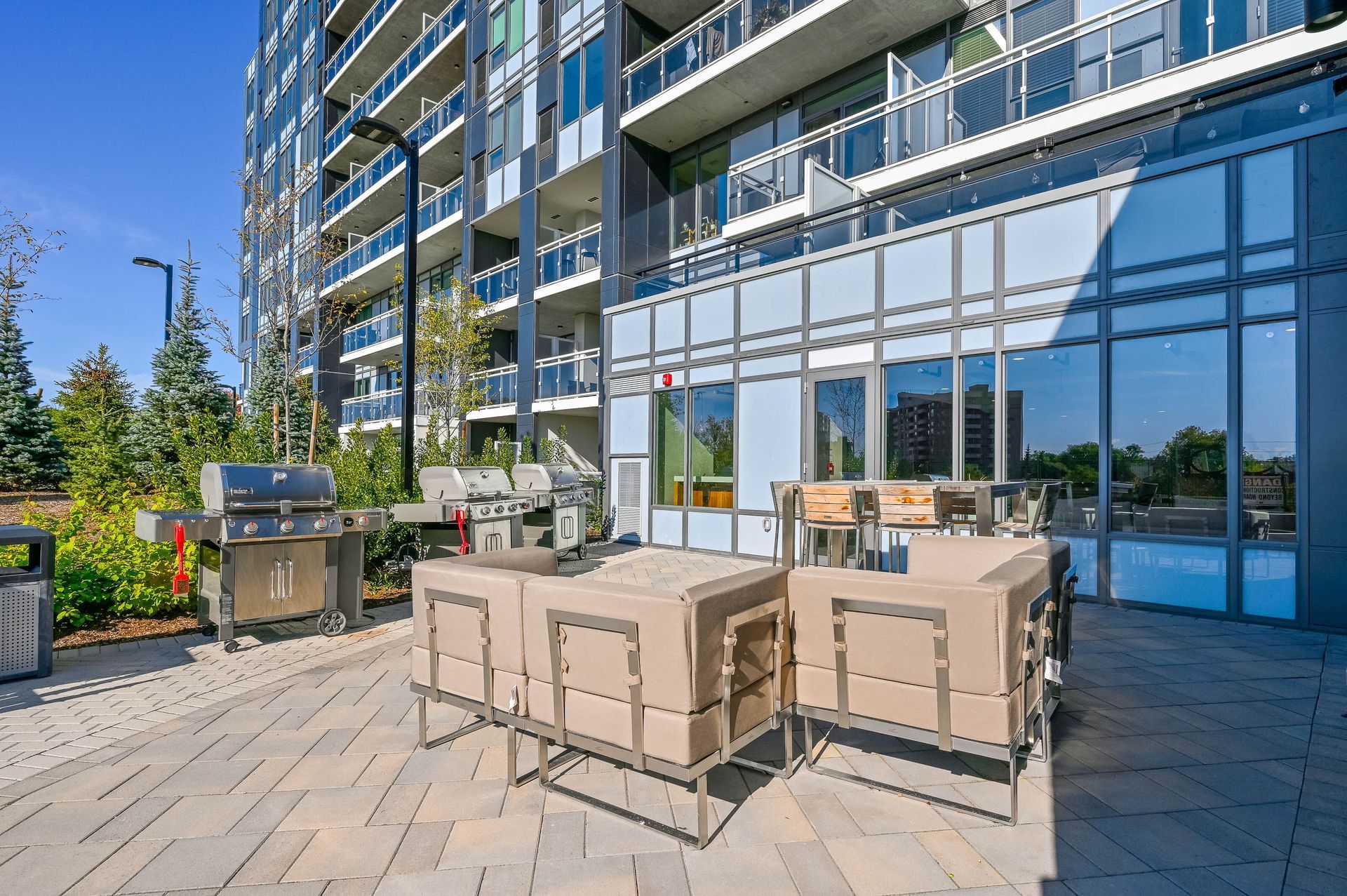 Outdoor patio with beige seating, grill, and dining table beside a modern glass apartment building.