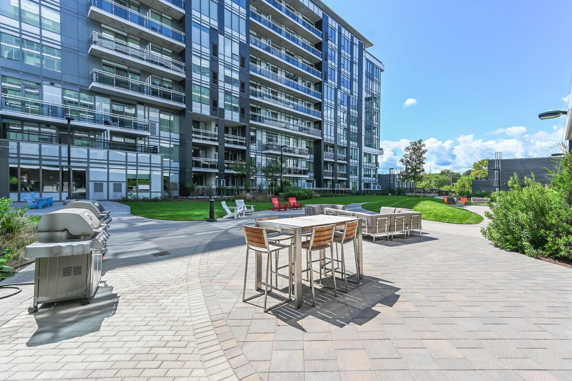 Modern apartment courtyard with outdoor dining tables, grill, and landscaped patio under a clear sky