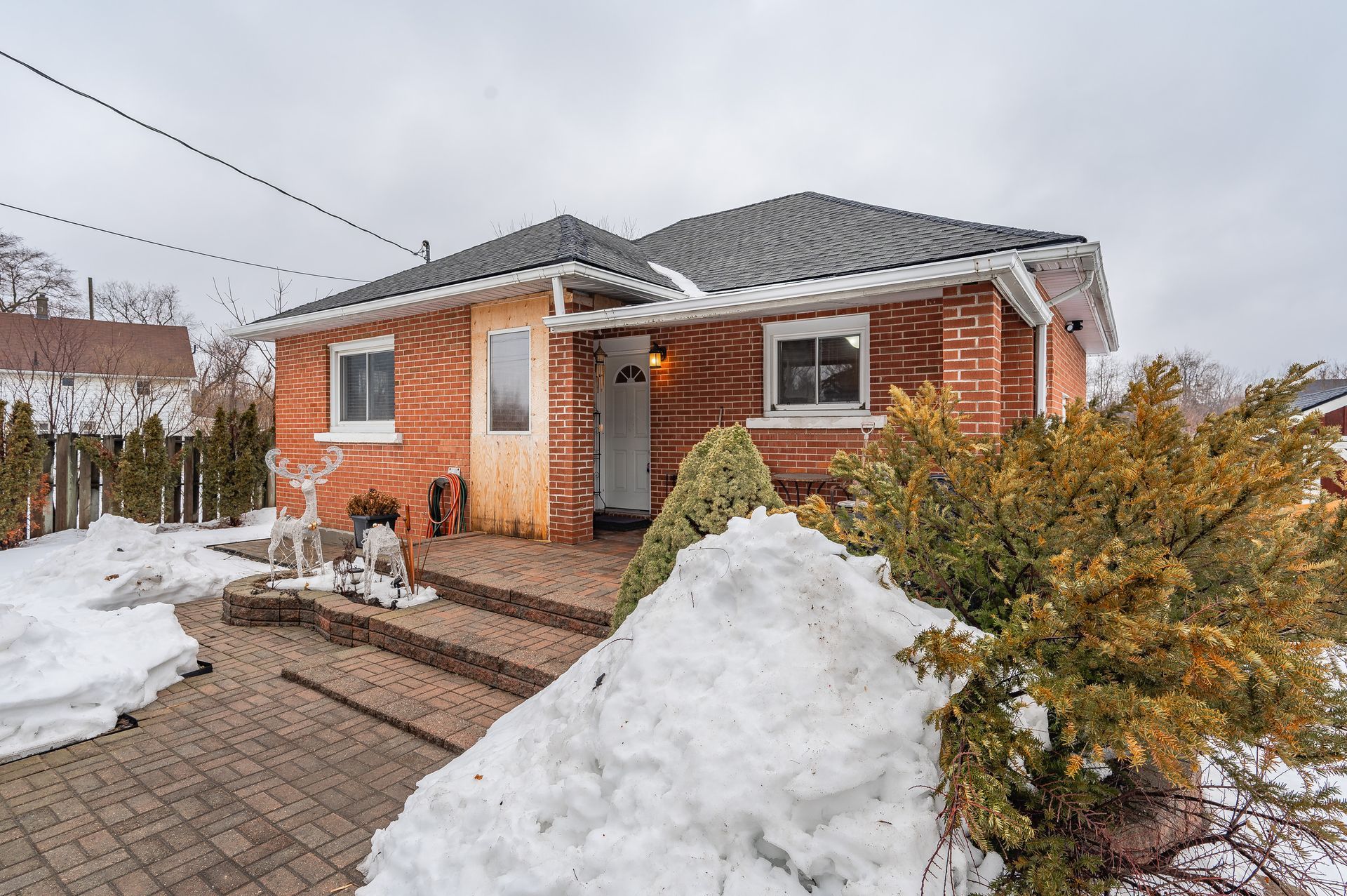 Snowy front yard with a small brick house, wooden walkway, and evergreen shrubs