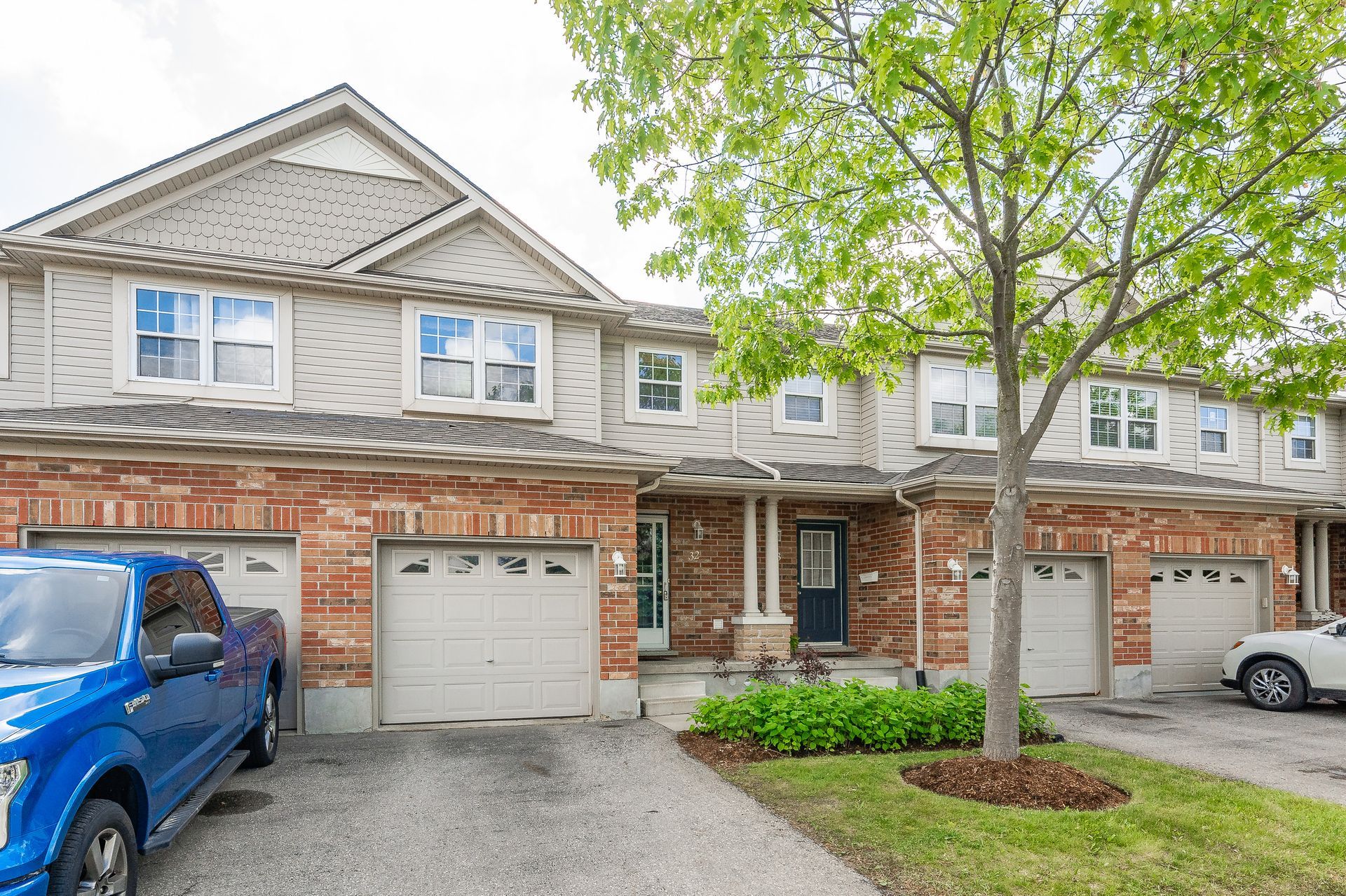 Townhouse row with brick and beige siding, garage doors, parked cars, and a tree in front