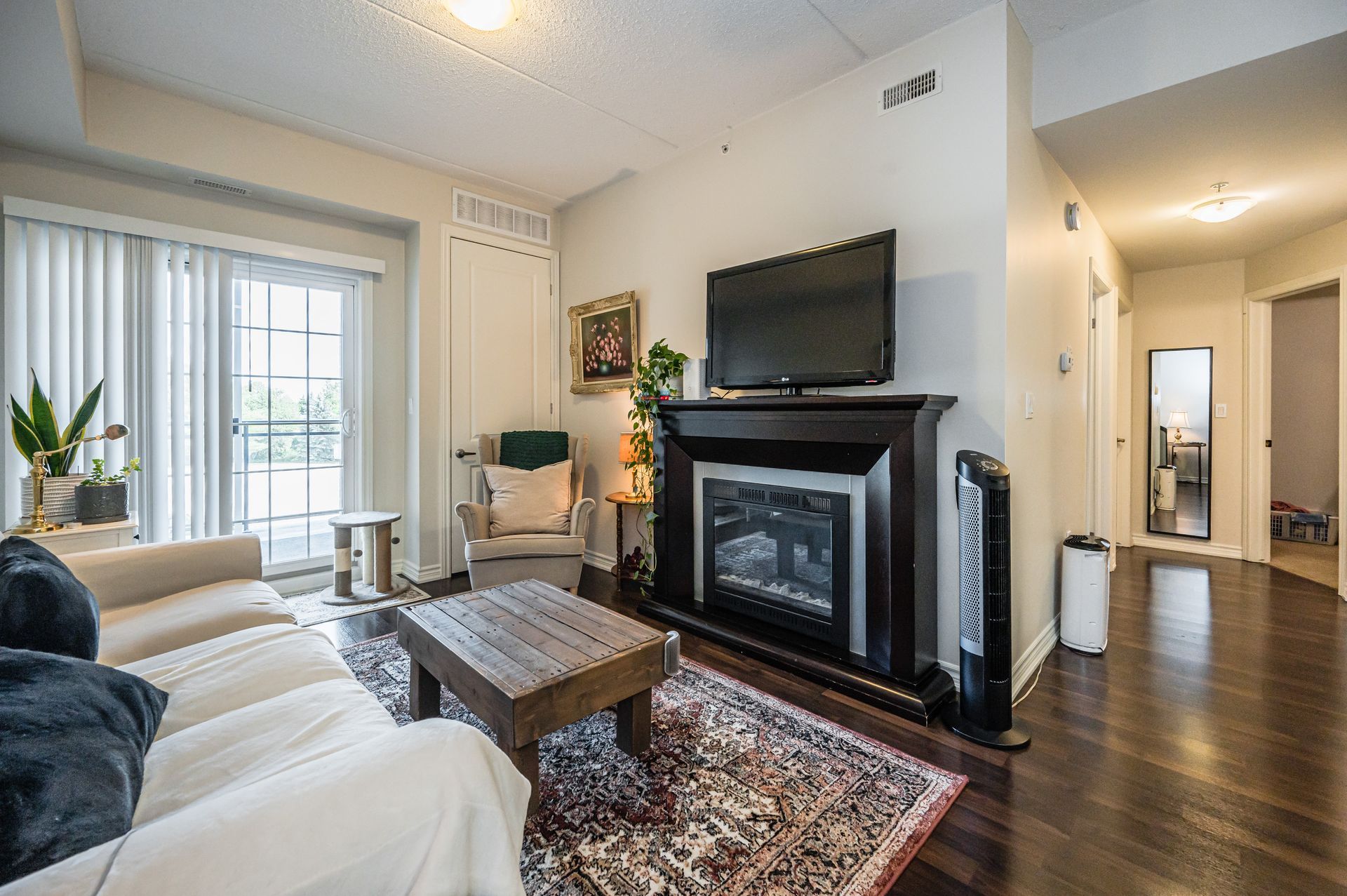 Bright living room with white sofa, fireplace, wall-mounted TV, hardwood floors, and balcony doors