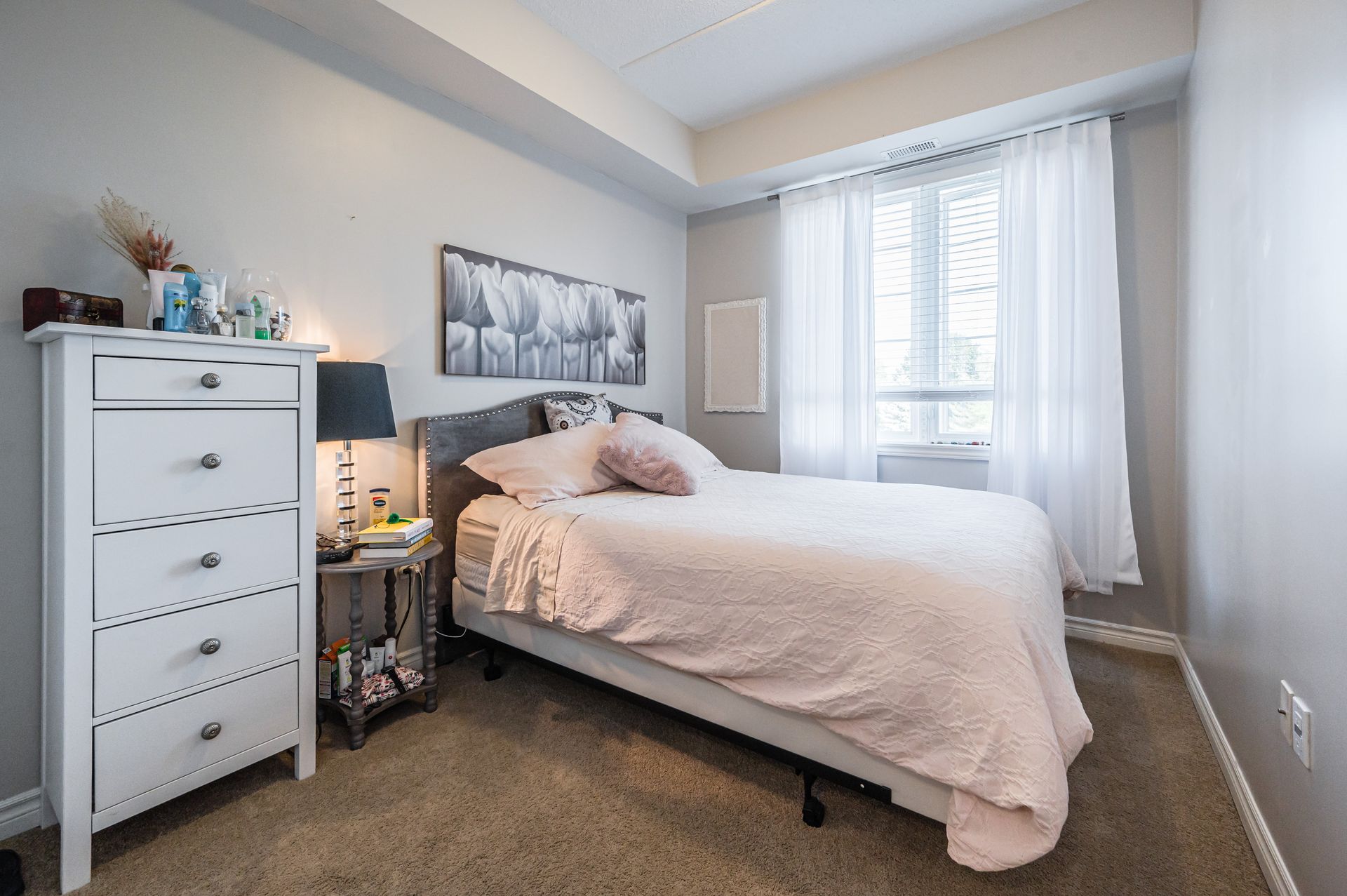 Bright bedroom with a bed, dresser, nightstand lamp, and window with sheer curtains