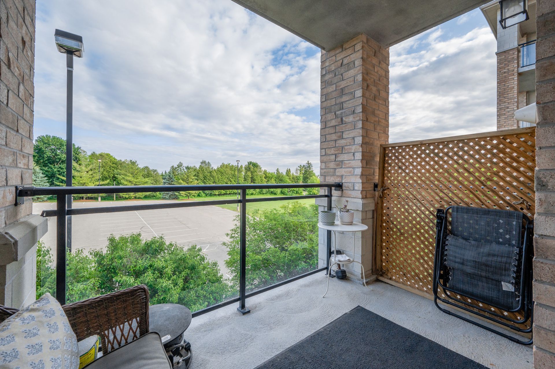 Covered balcony with black railing, wicker chairs, and a view of trees and a cloudy sky.