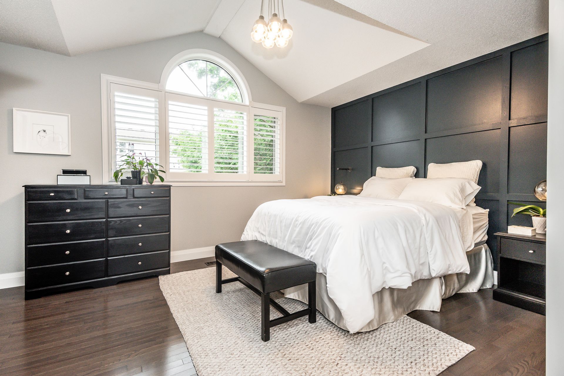 Bright bedroom with dark paneled wall, white bed, black dresser, arched window, and bench on a rug