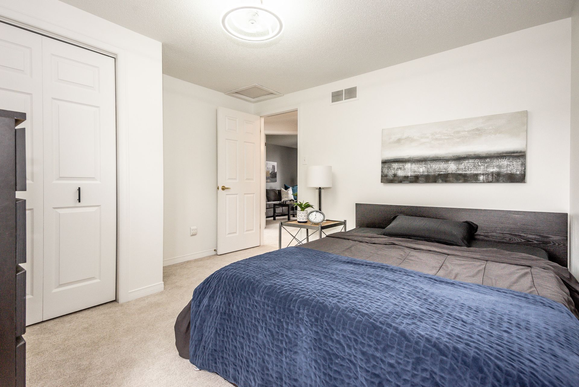 Bright bedroom with a blue bedspread, white walls, carpet, and a closet door open to a desk area