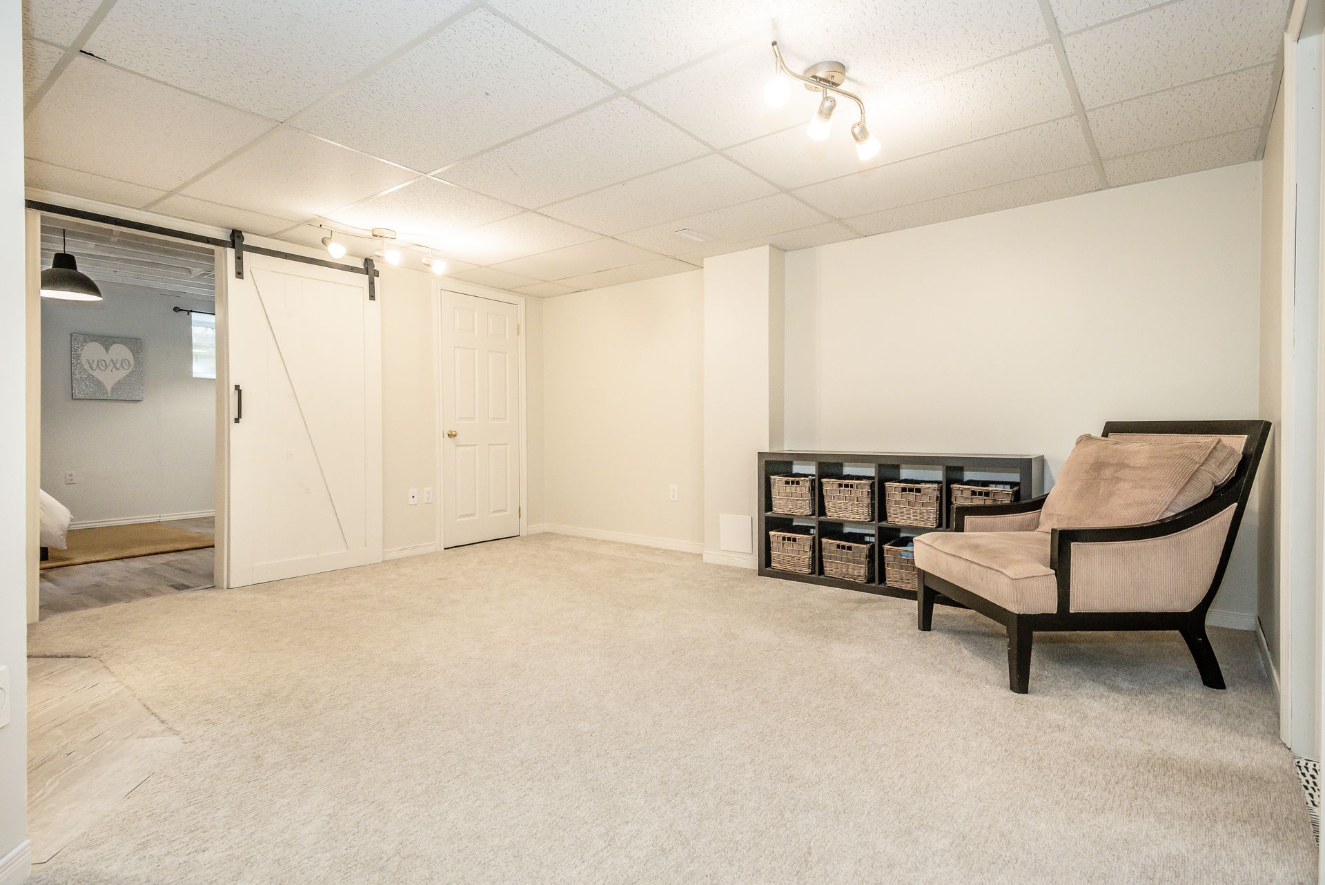 Empty carpeted basement room with white walls, ceiling lights, and a chair near storage bins