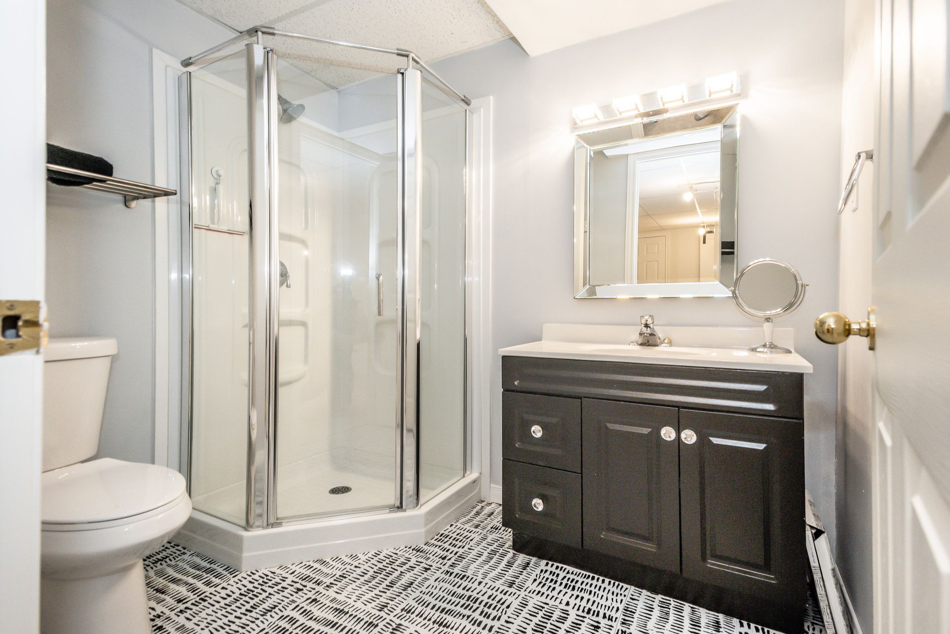 Bright white bathroom with glass shower, patterned floor, vanity, mirror, and toilet.