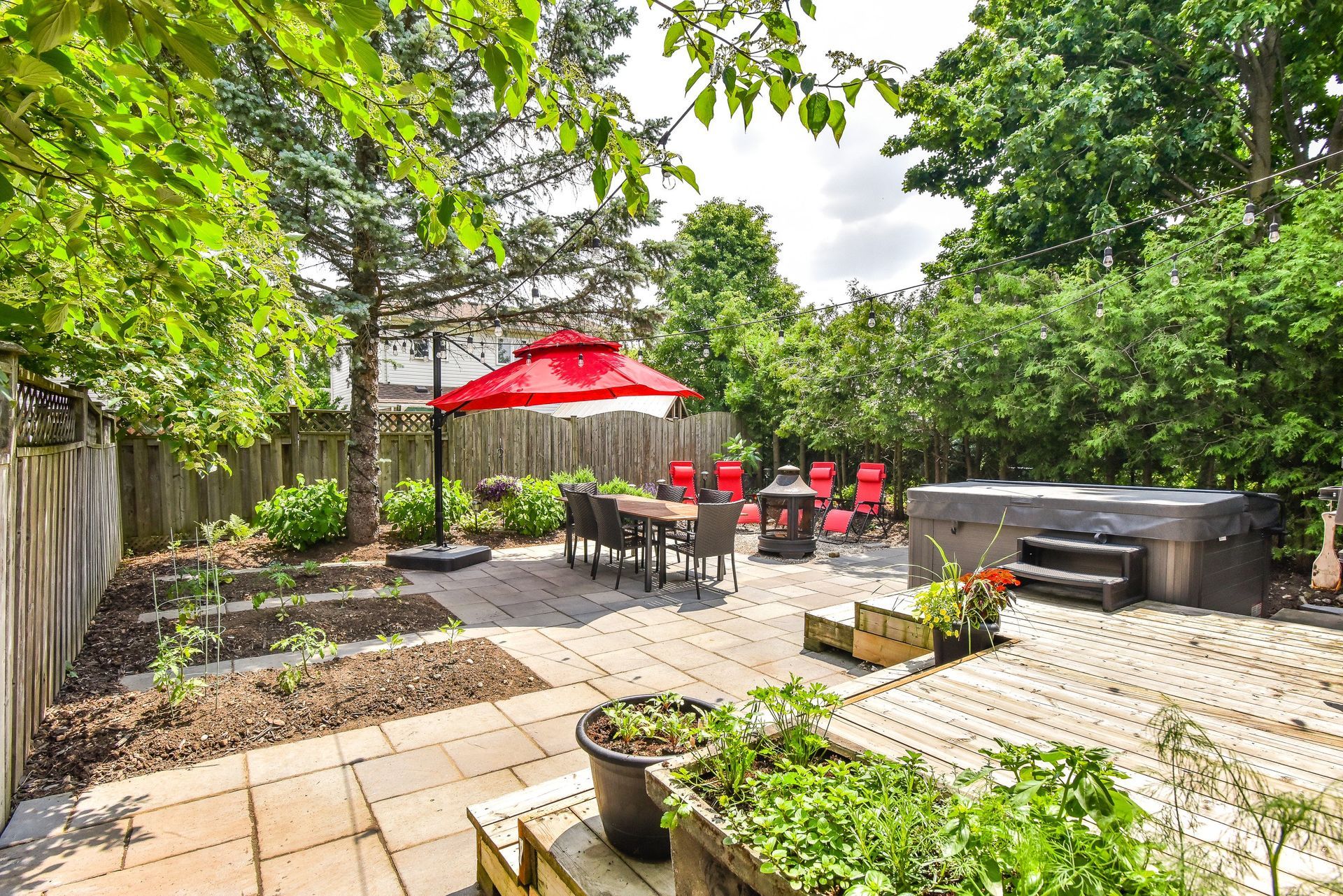 Backyard patio with red umbrella, table and chairs, potted plants, and a hot tub amid lush greenery