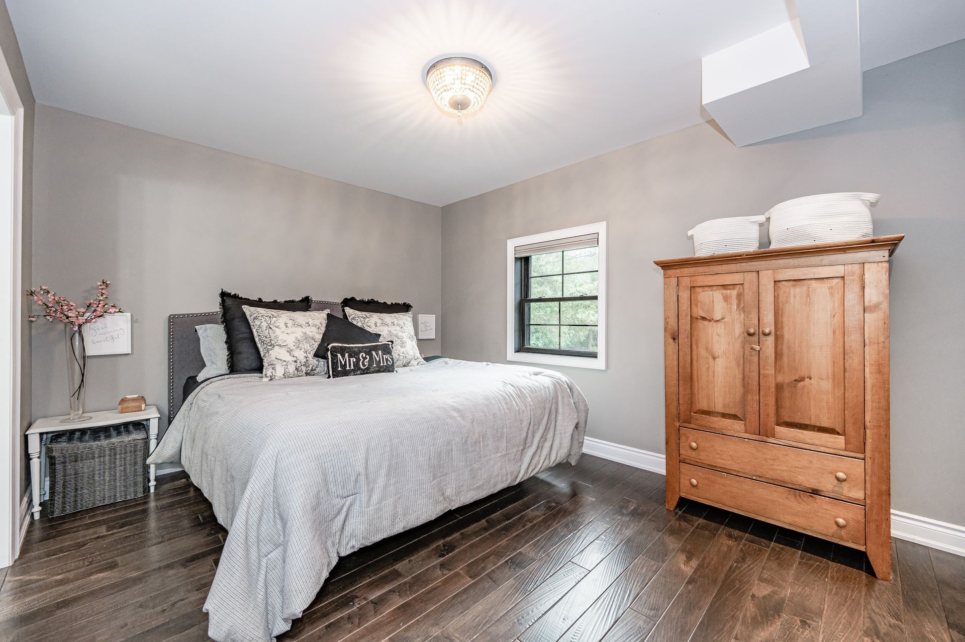 Bright bedroom with a bed, wooden dresser, small window, and dark wood floors.